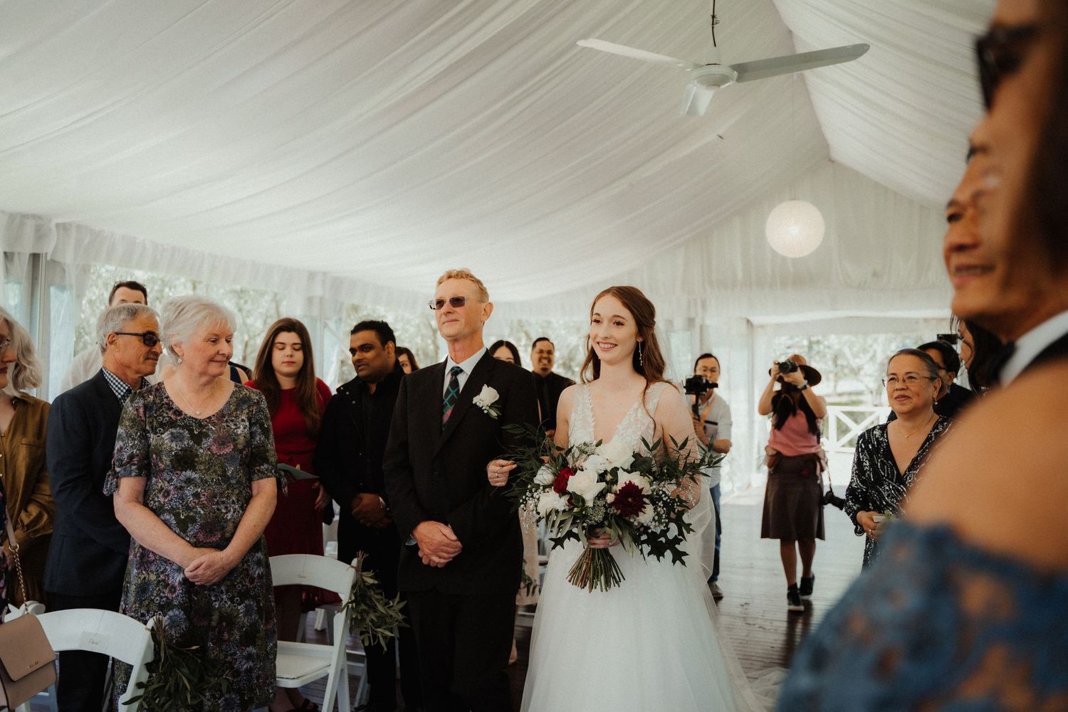 Bride in a white gown with a floral bouquet walks down the aisle toward the groom as guests watch at an indoor wedding ceremony.