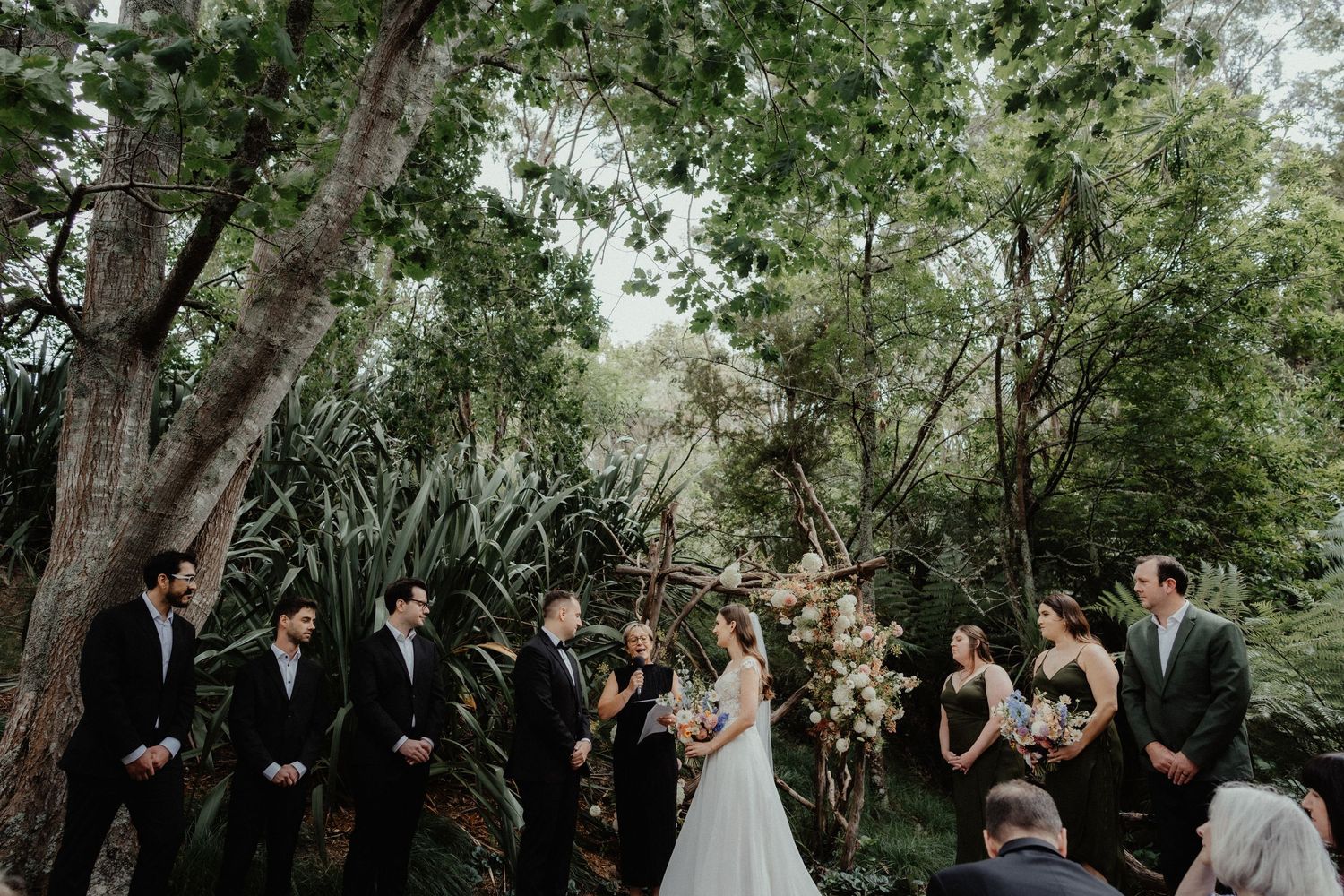 A bride and groom stand before a floral arch with their wedding party during a woodland ceremony beneath tall trees.