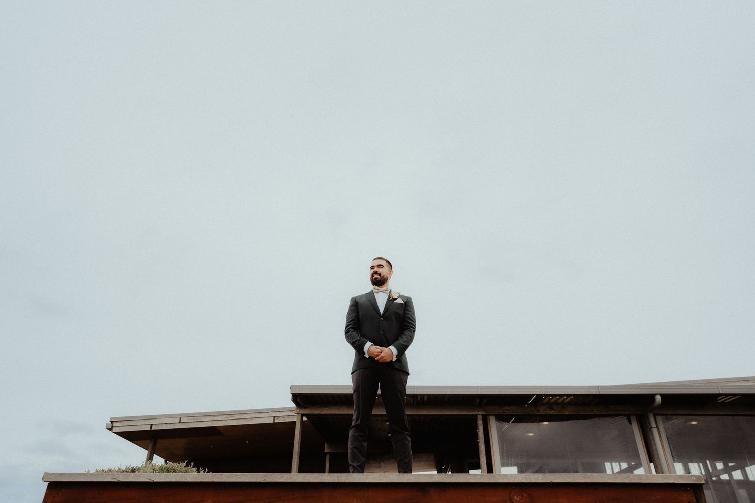 Groom in a dark suit stands alone on a rooftop ledge looking into the distance beneath a pale overcast sky.