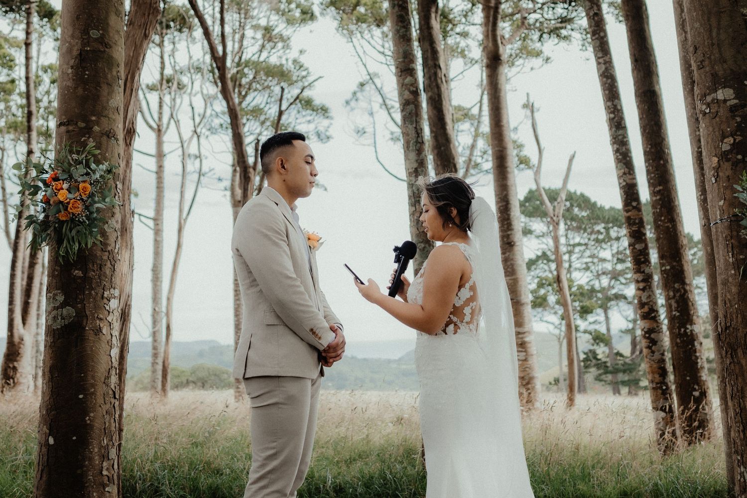 A groom in a beige suit listens as the bride in a white gown reads her vows among tall trees with a floral arrangement nearby.