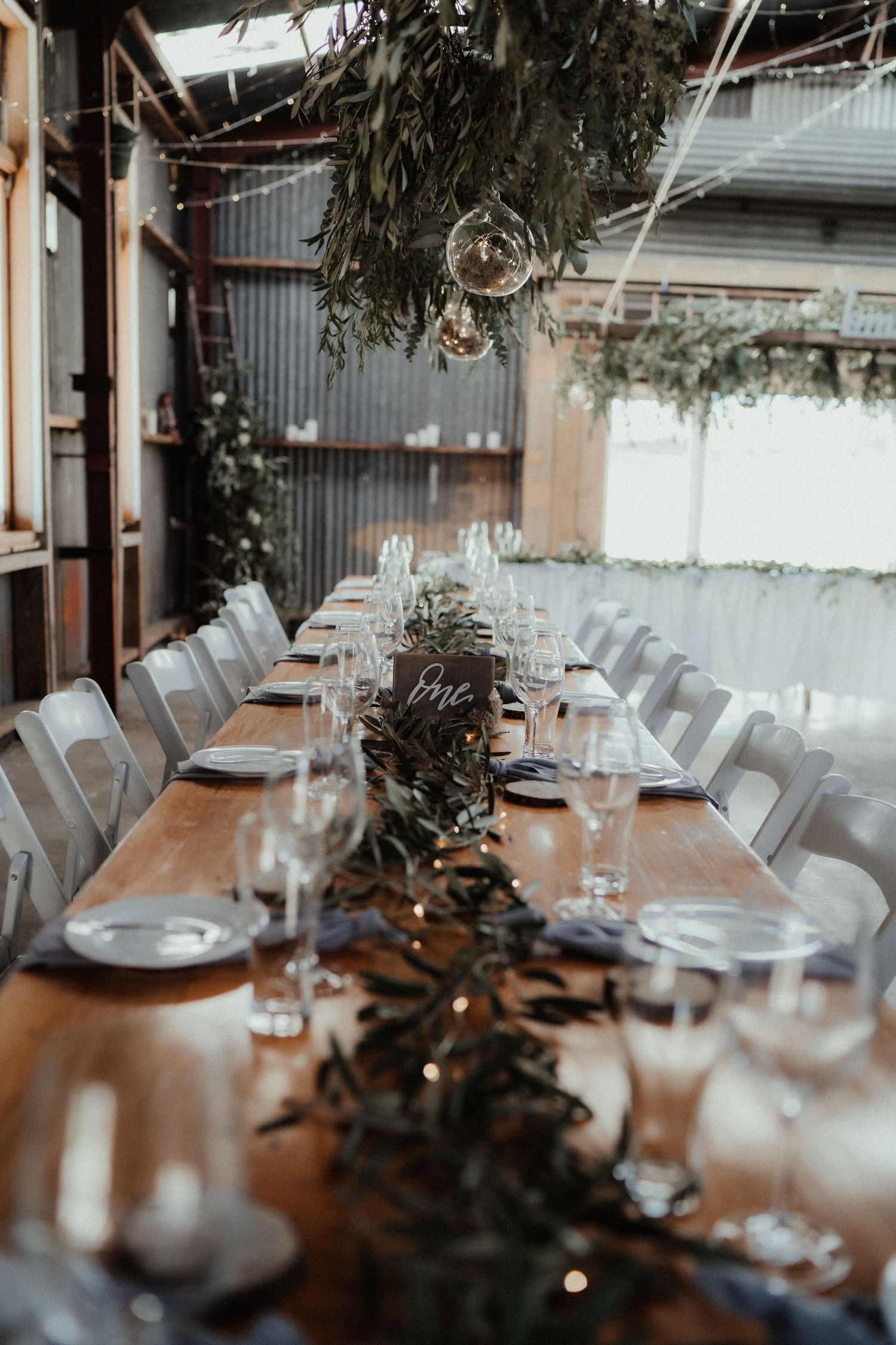 Long rustic wooden reception table styled with greenery runners, glassware and hanging foliage in a barn wedding venue.