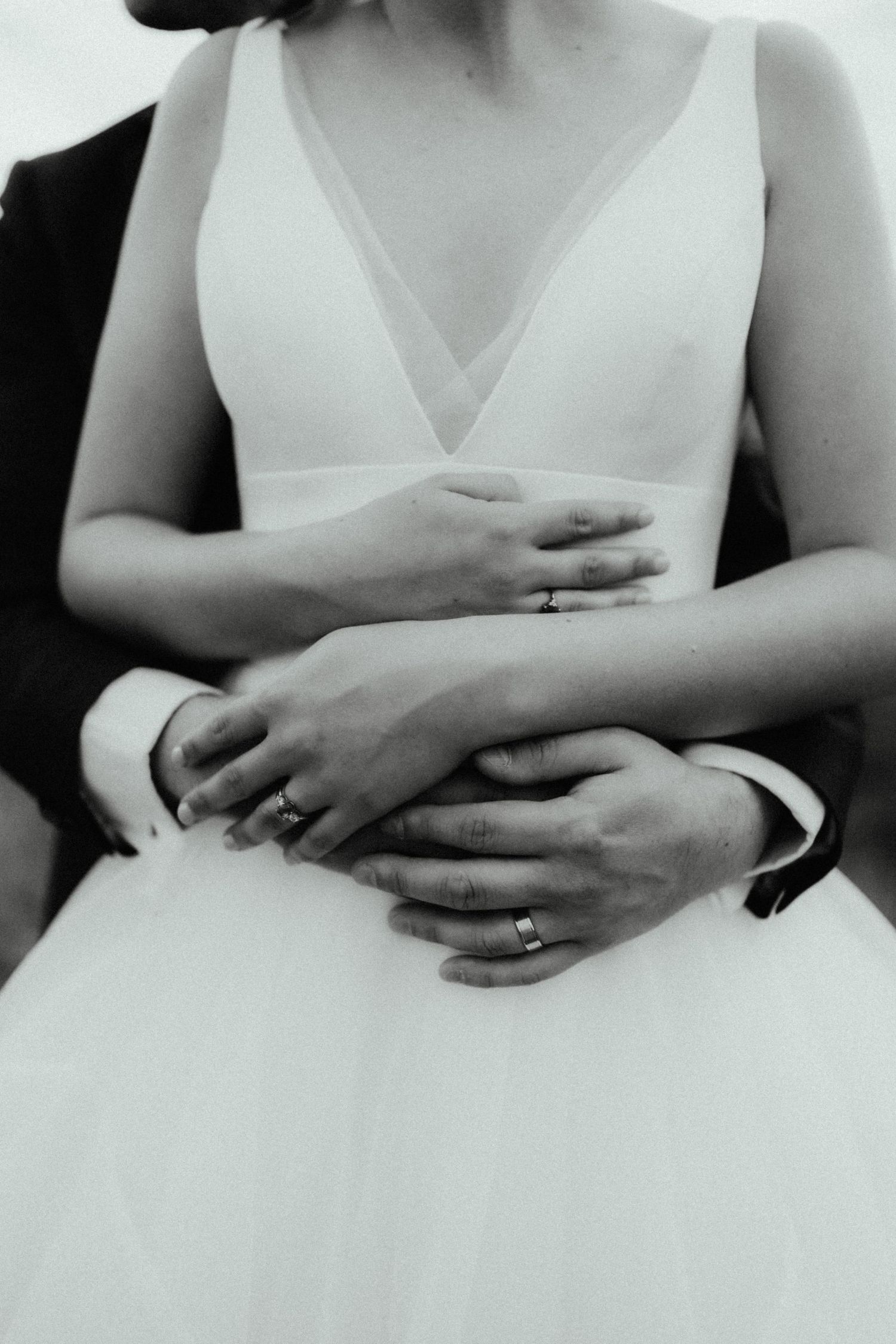 Close-up black and white photo of a bride and groom's hands with wedding rings embracing over a tulle wedding gown.
