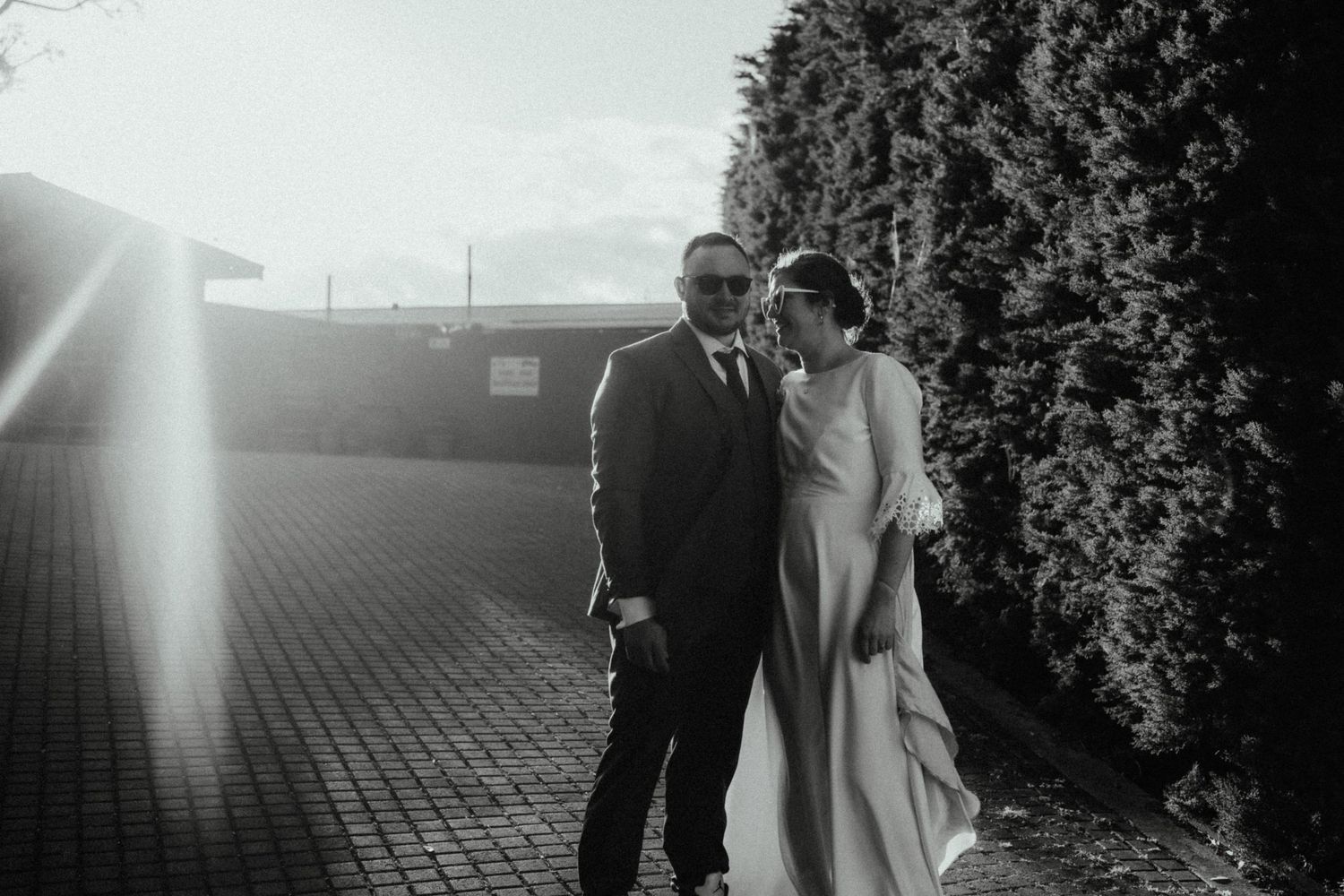 Black and white wedding portrait of a couple in sunglasses and formal wear standing together beside tall hedges in sunlight.
