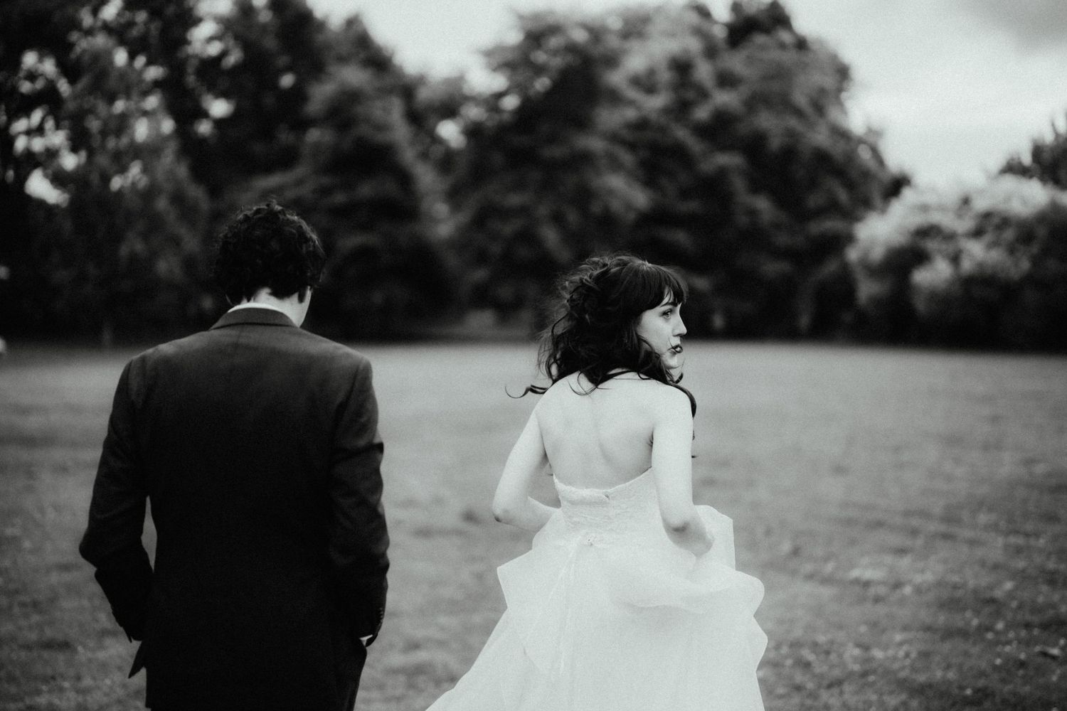Black and white photo of a bride in a flowing tulle gown glancing back as she walks across a park lawn with her groom.