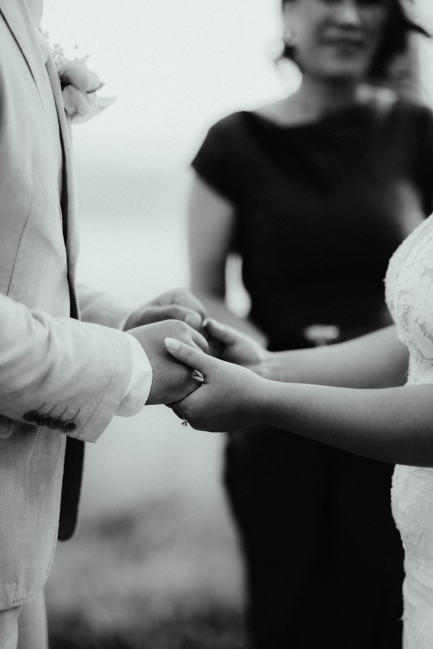 Black and white close-up of the couple's joined hands during the ceremony as the celebrant smiles behind.