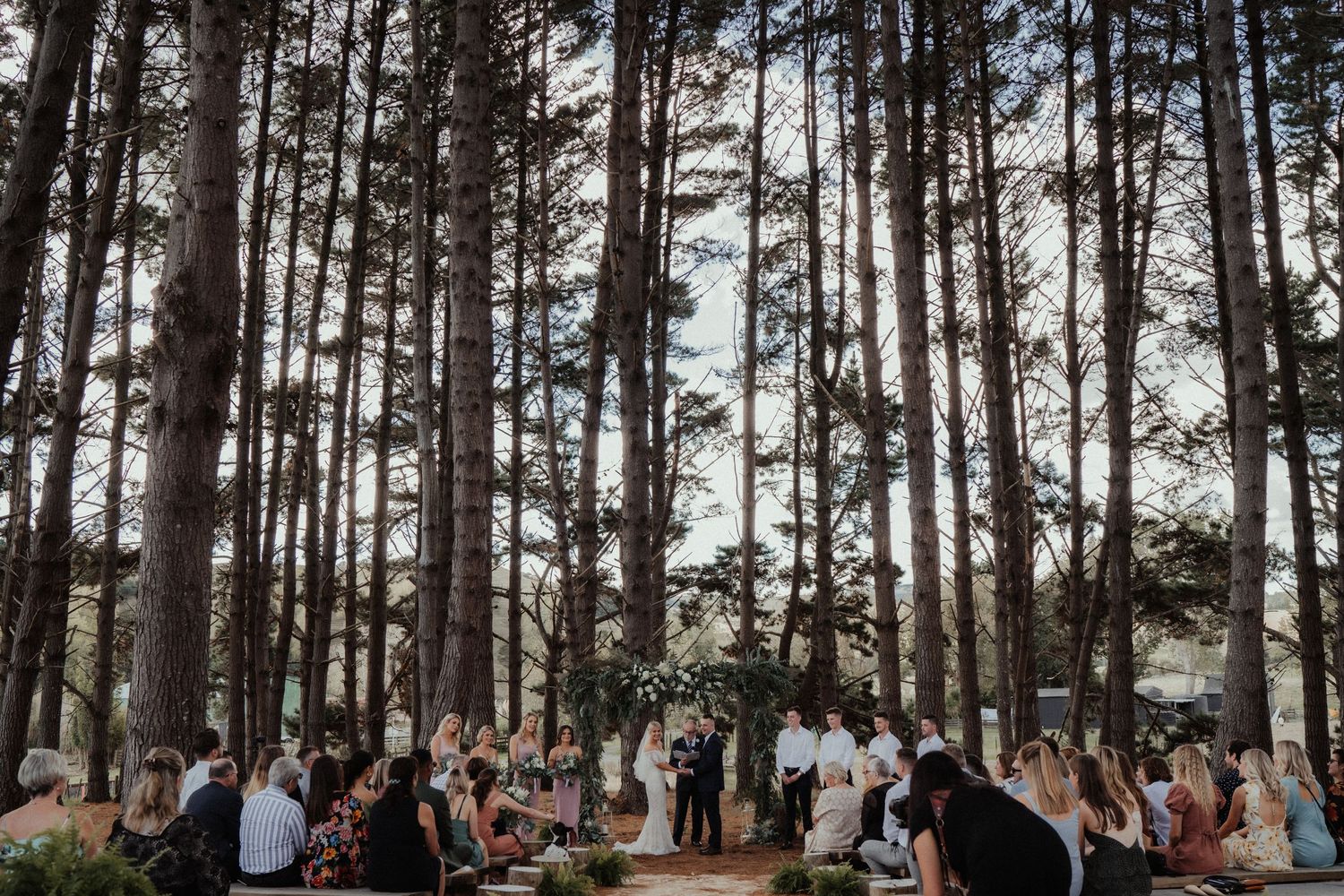 Wedding guests sit among tall pine trees as a bride and groom stand at a floral altar during an outdoor forest ceremony.