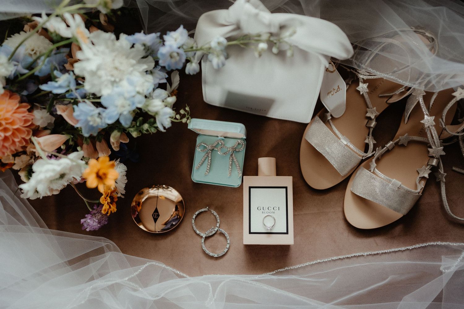 Wedding flat lay featuring a pastel bouquet, strappy sandals, rings, perfume, a handbag and a small jewelry box on a wooden surface.