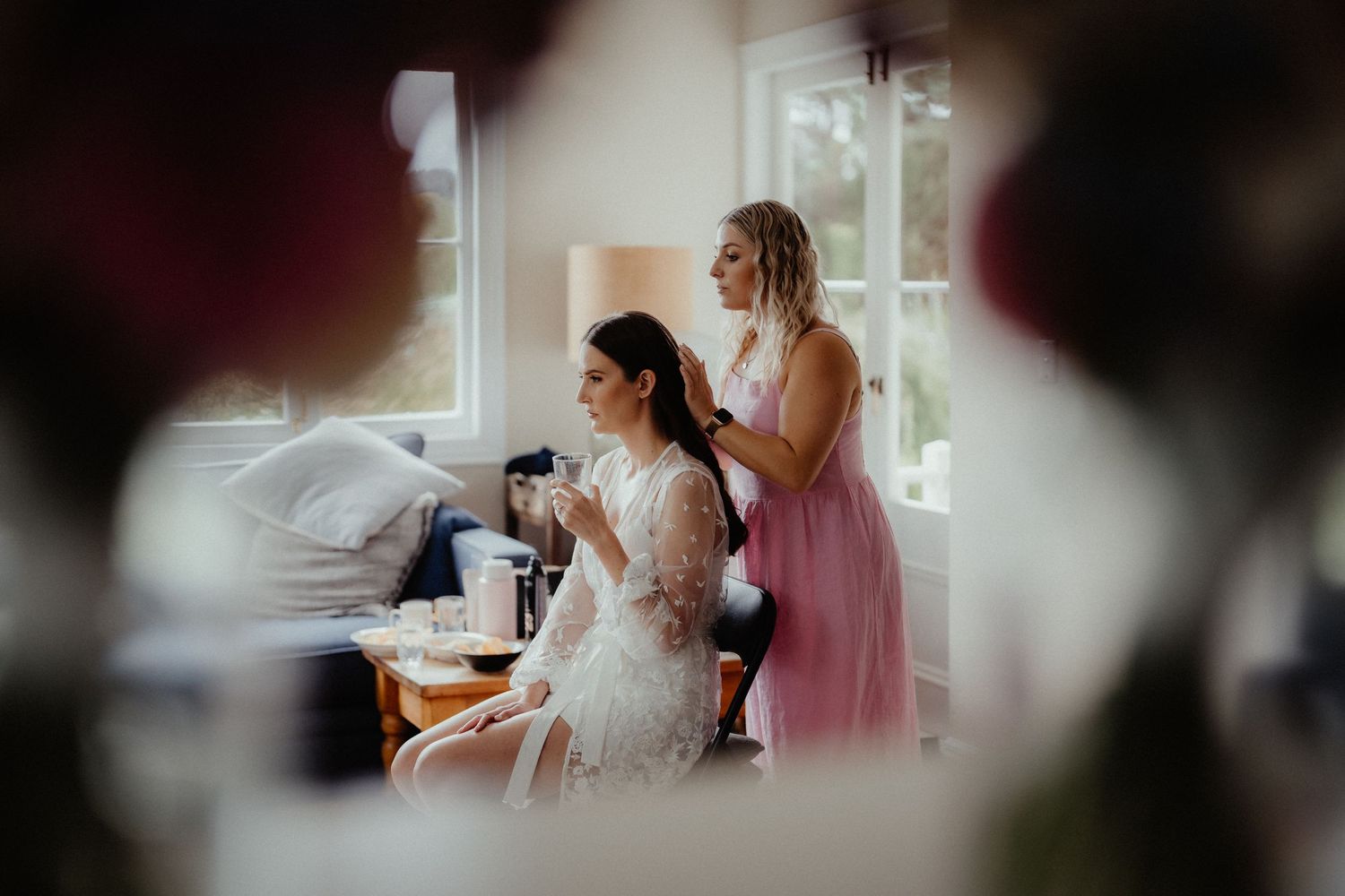 Bridesmaid in a pink dress styles the bride's hair as the bride in a lace robe sips a drink in a sunlit room.