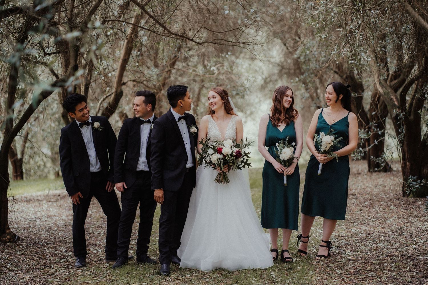 Bride stands with groomsmen in black tuxedos and bridesmaids in teal dresses in an olive grove.