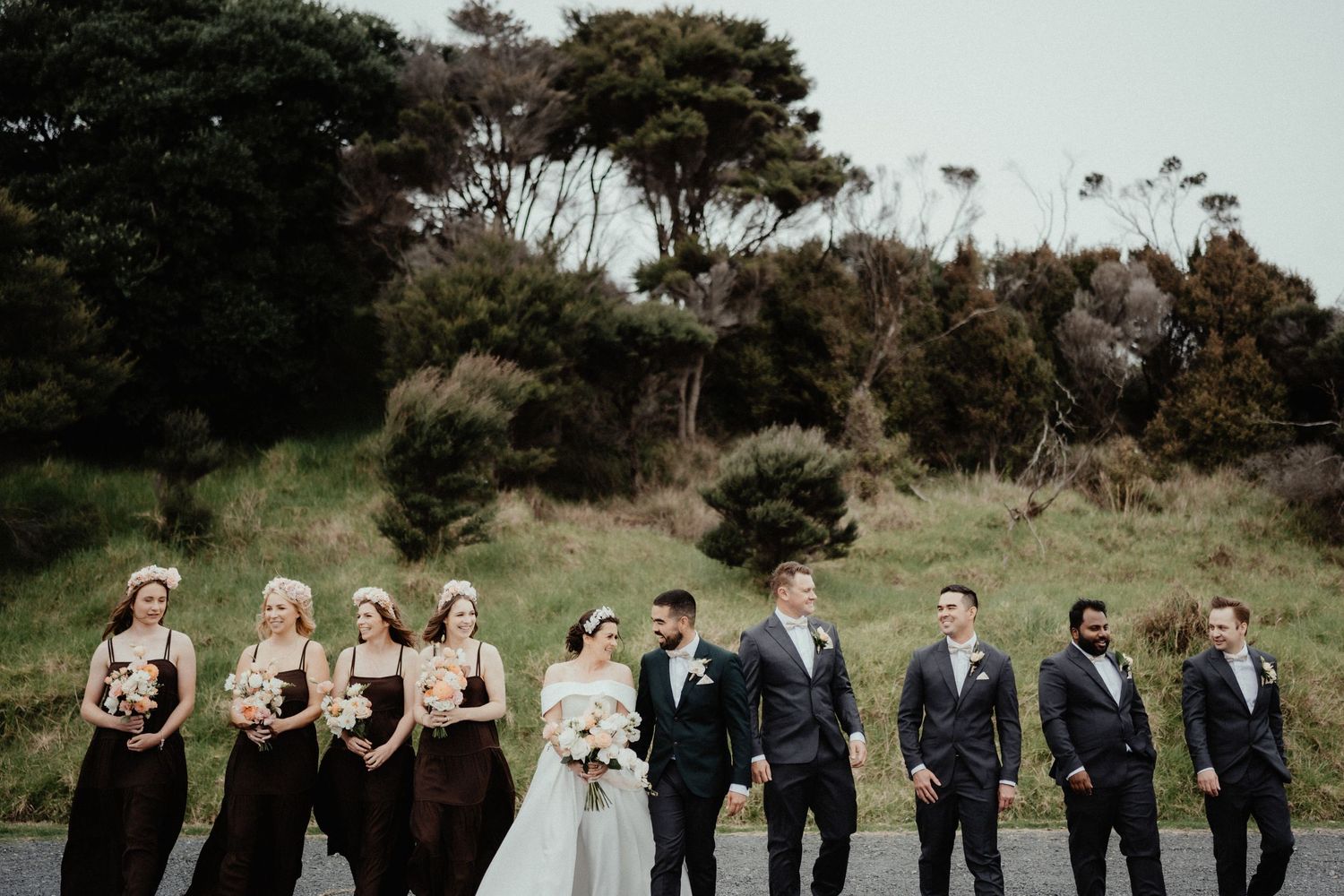 Bridal party of bridesmaids in brown dresses and groomsmen in gray suits walks with the couple past a grassy hillside.