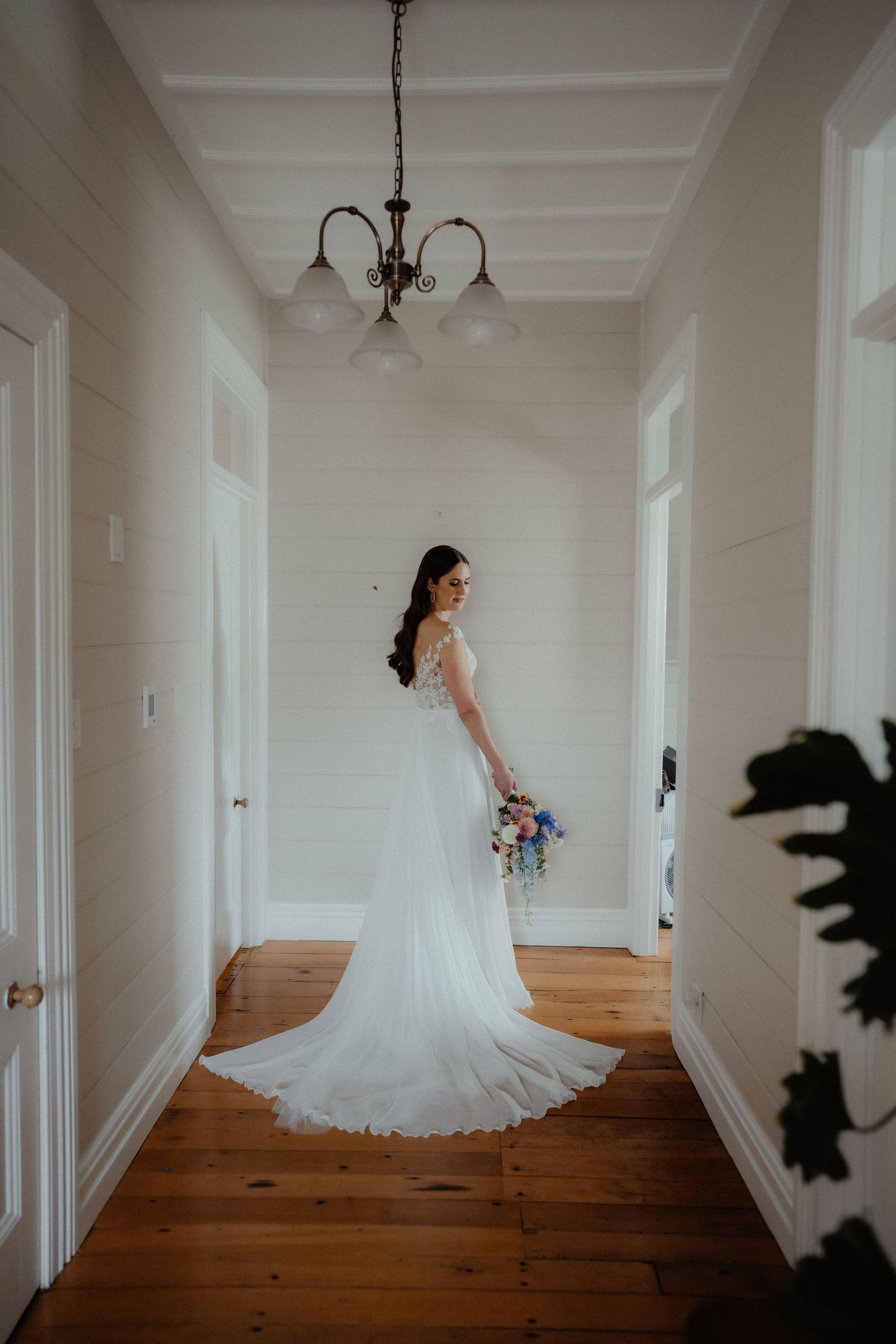 Bride in a flowing white wedding gown holding a bouquet while standing in a bright hallway with wooden floors.