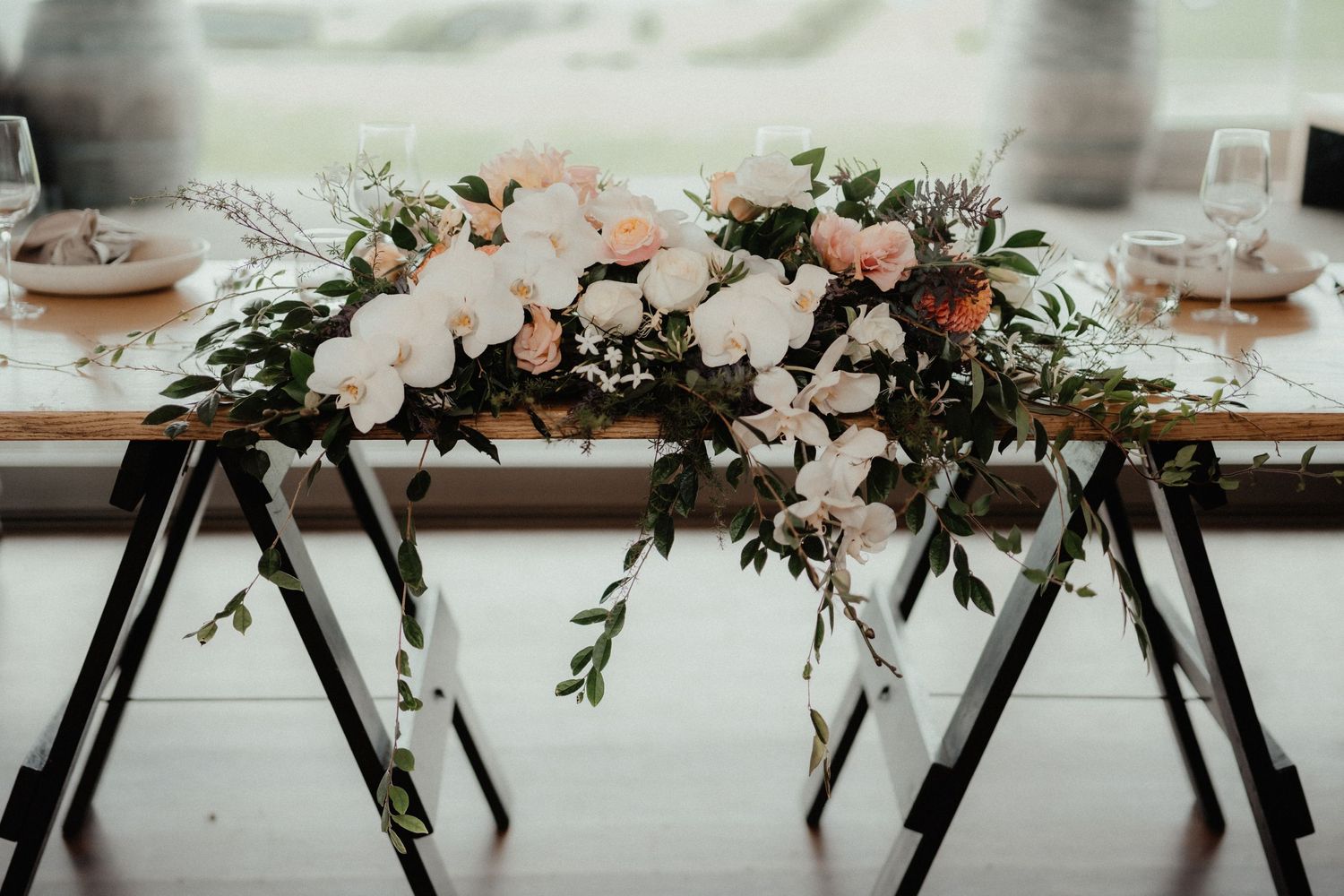 Bridal table decorated with a lush floral garland of white orchids, pink blooms and greenery on a wooden trestle table.