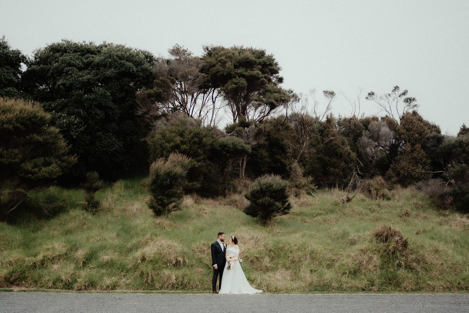 Bride and groom stand hand in hand on a quiet road in front of a grassy hillside and windswept coastal trees.