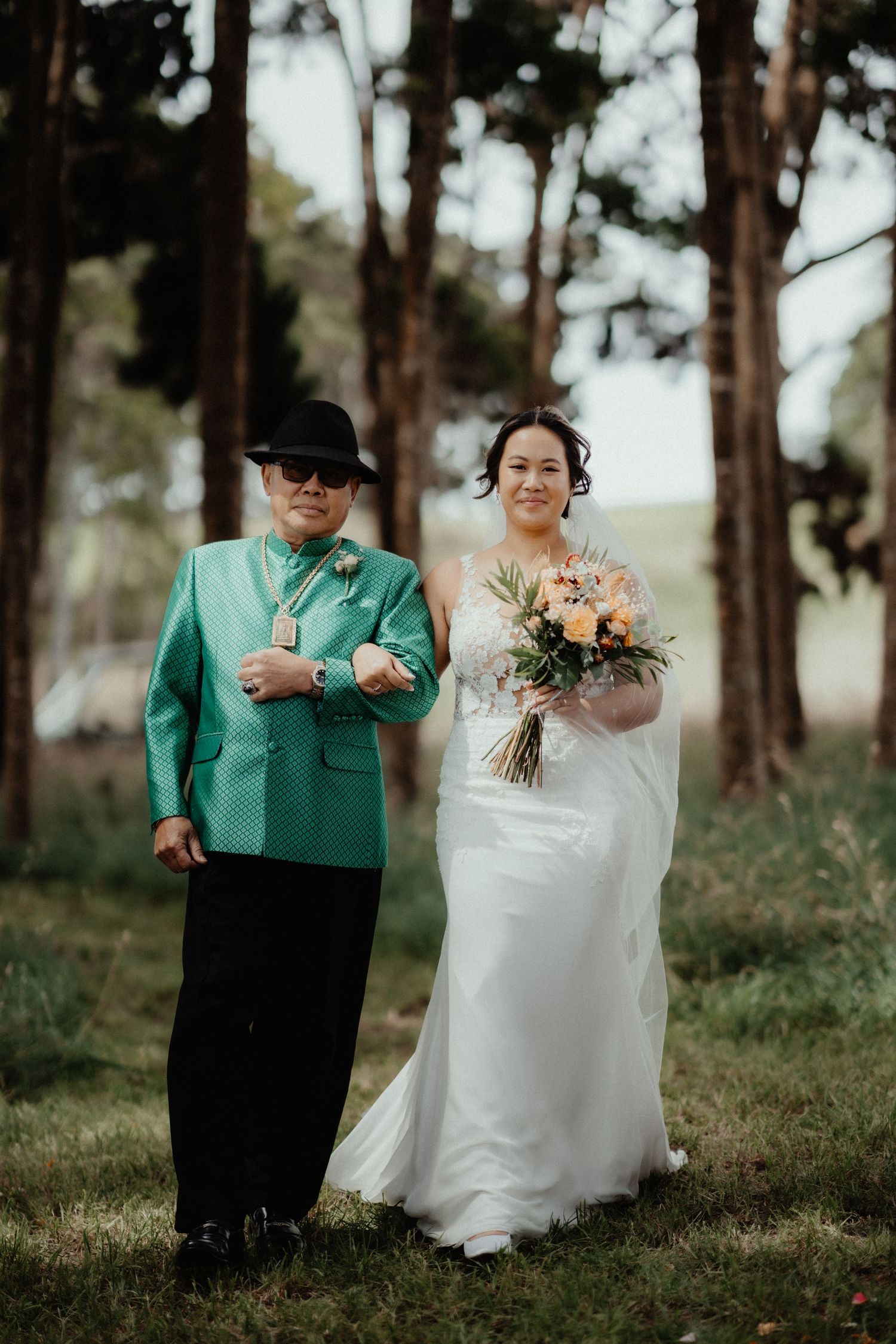 Bride carrying a peach bouquet is escorted down a tree-lined aisle by a man in a green jacket and black hat.