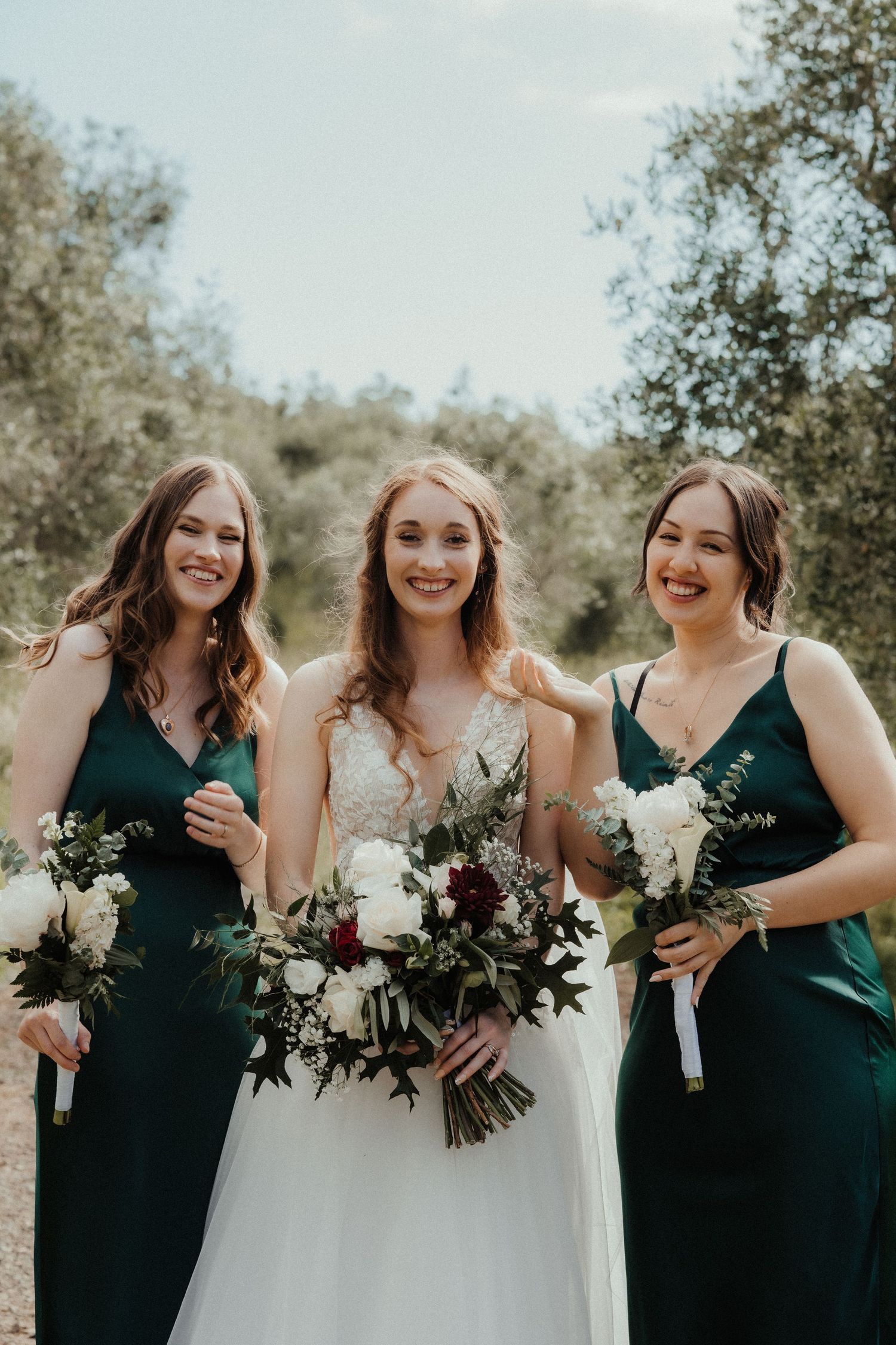 Smiling bride with long wavy hair stands between two bridesmaids in green dresses, all holding white and burgundy floral bouquets.