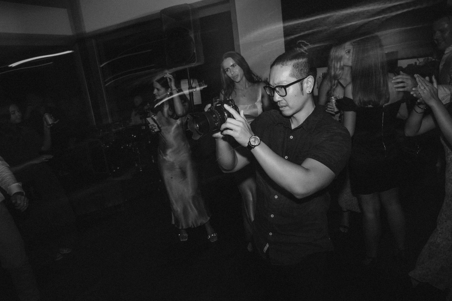 Black and white photo of a smiling man with glasses holding a camera as guests dance at a wedding reception.
