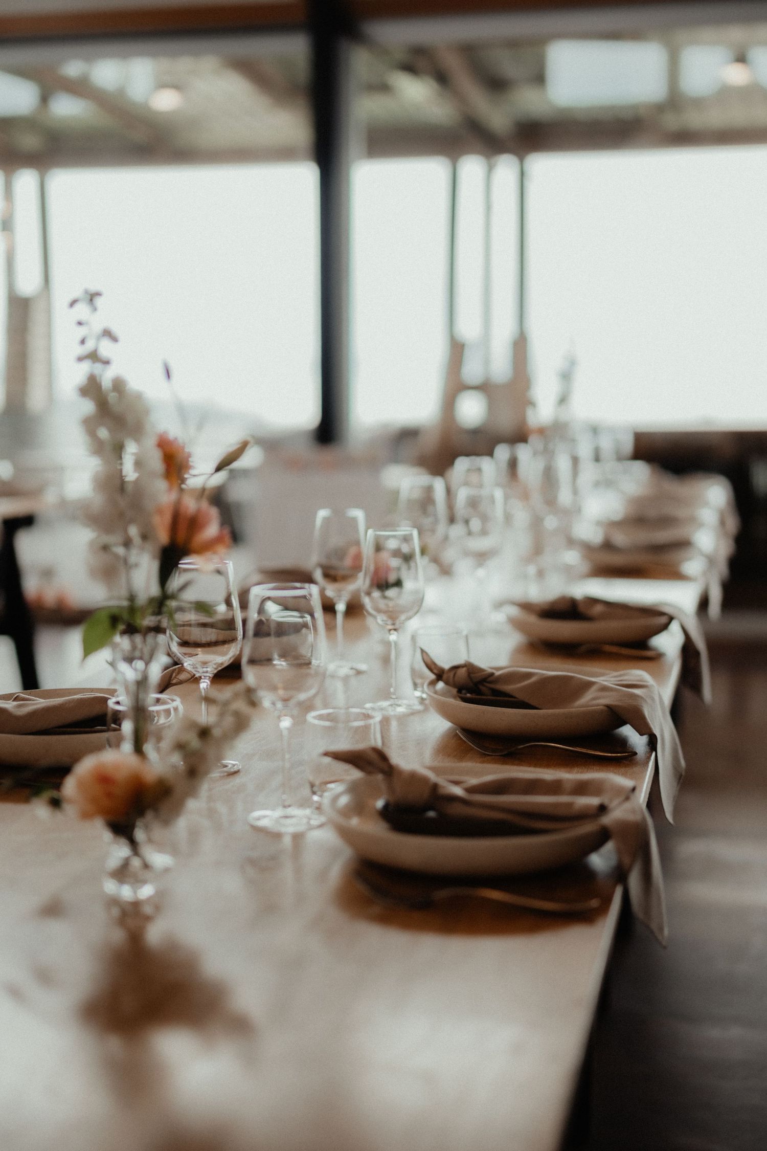 Elegant long wedding table set with wine glasses, linen napkins, plates and delicate peach floral arrangements by a large window.