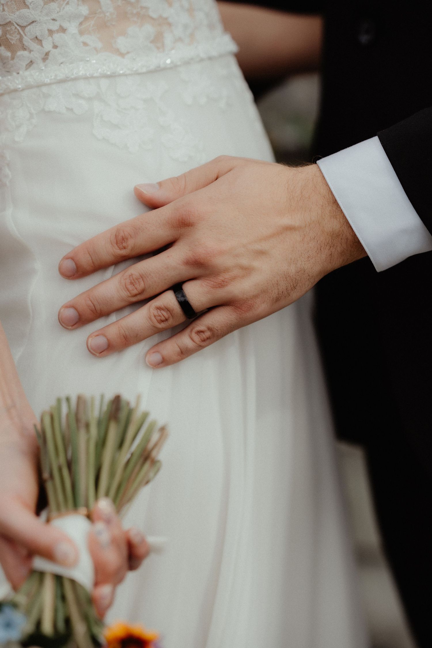Close-up of a groom's hand with a black wedding band resting on the bride's lace dress as she holds a bouquet.