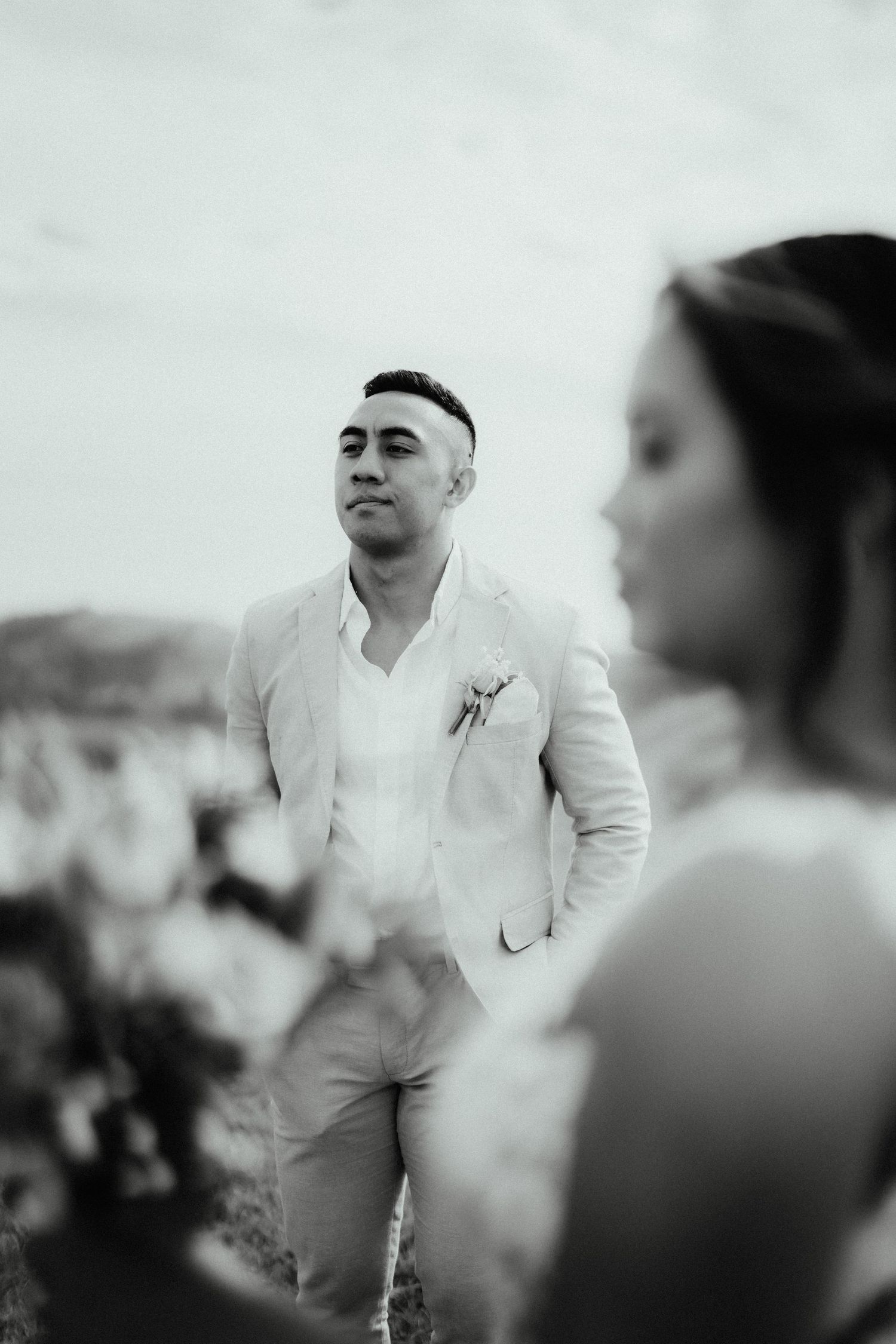 Black and white portrait of a groom in a linen suit with a boutonniere gazing into the distance as the bride is softly blurred in front.