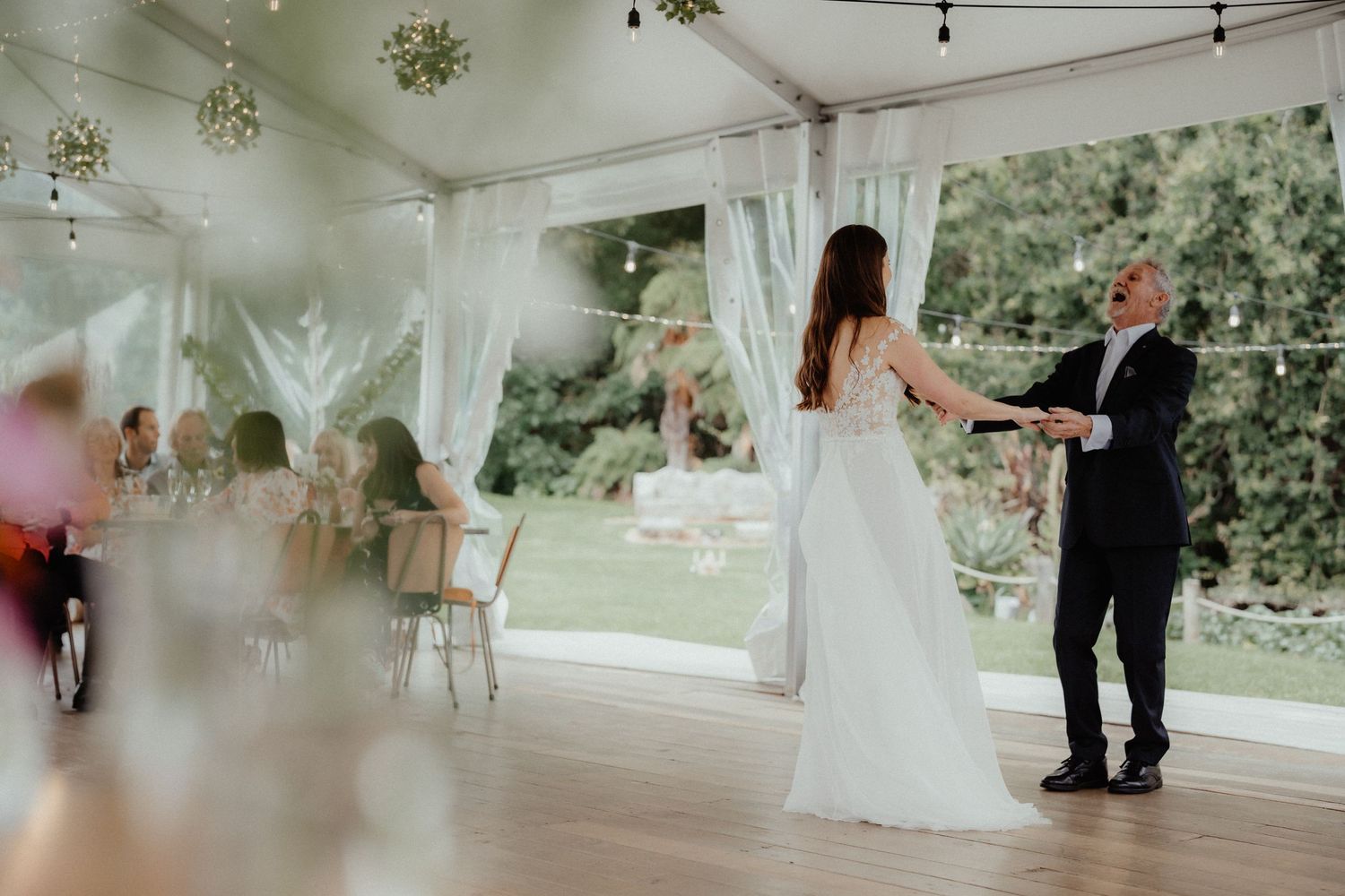 Bride in a flowing white gown dances with an older man on a wooden dance floor inside a white marquee with garden views.