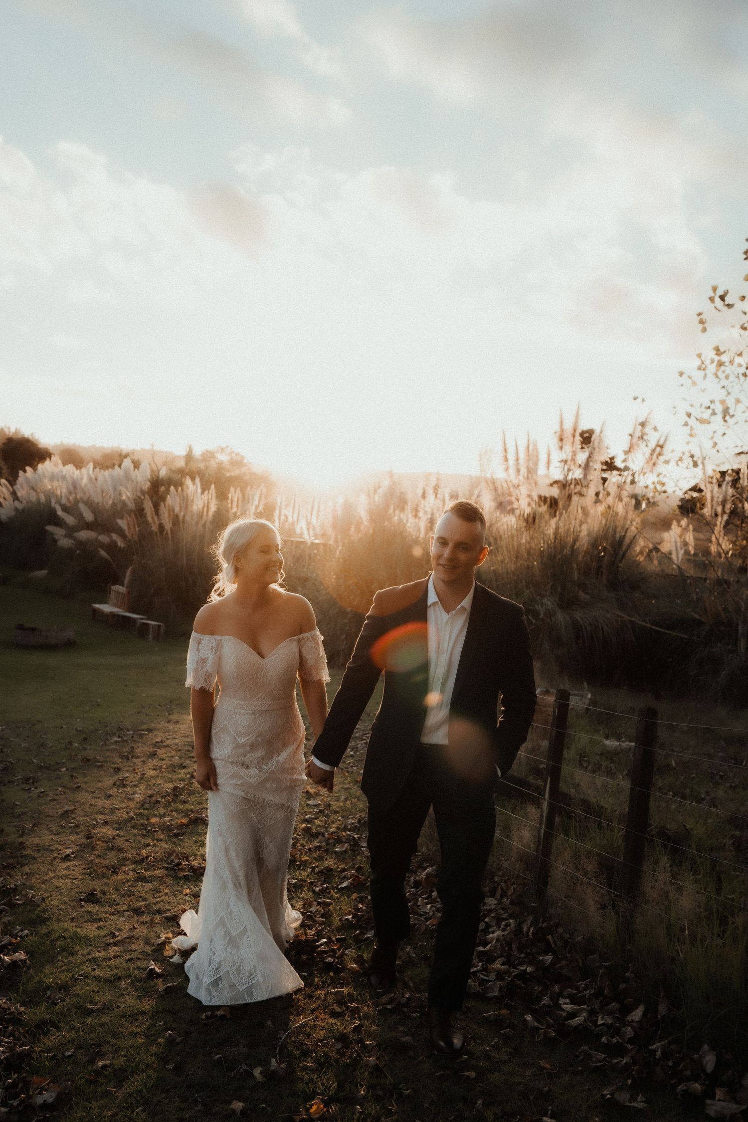 Bride in a lace gown and groom in a dark suit hold hands and walk through a golden-lit field at sunset with warm lens flare.