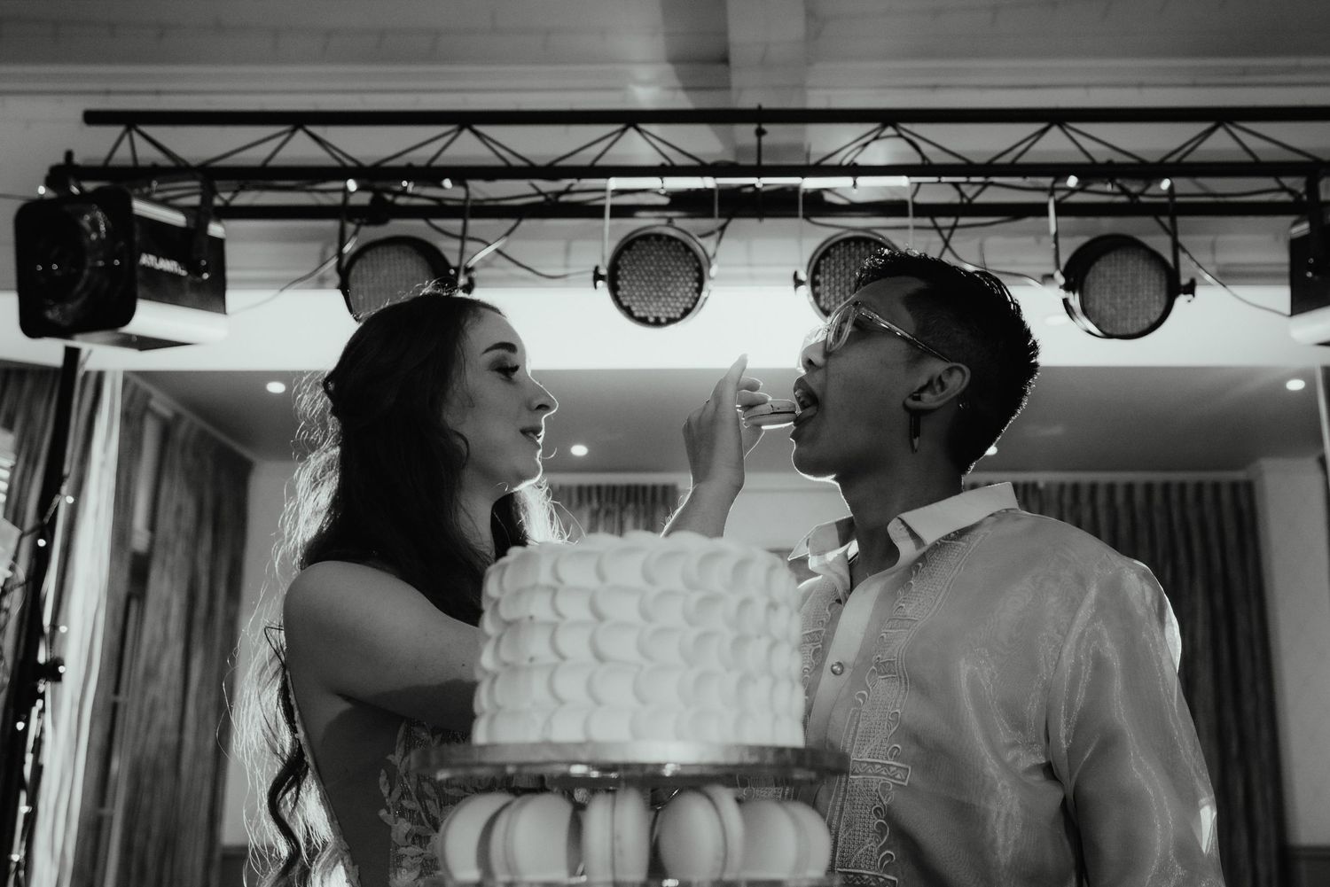 Black and white photo of a bride and groom playfully feeding each other cake beside a tiered wedding cake under stage lighting.