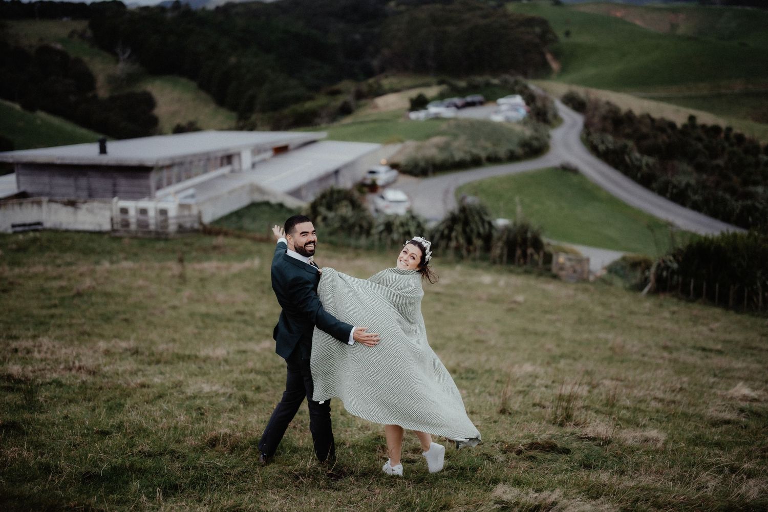 Groom twirls his bride in a flowing pale dress on a grassy hillside with farm buildings and a winding road below.