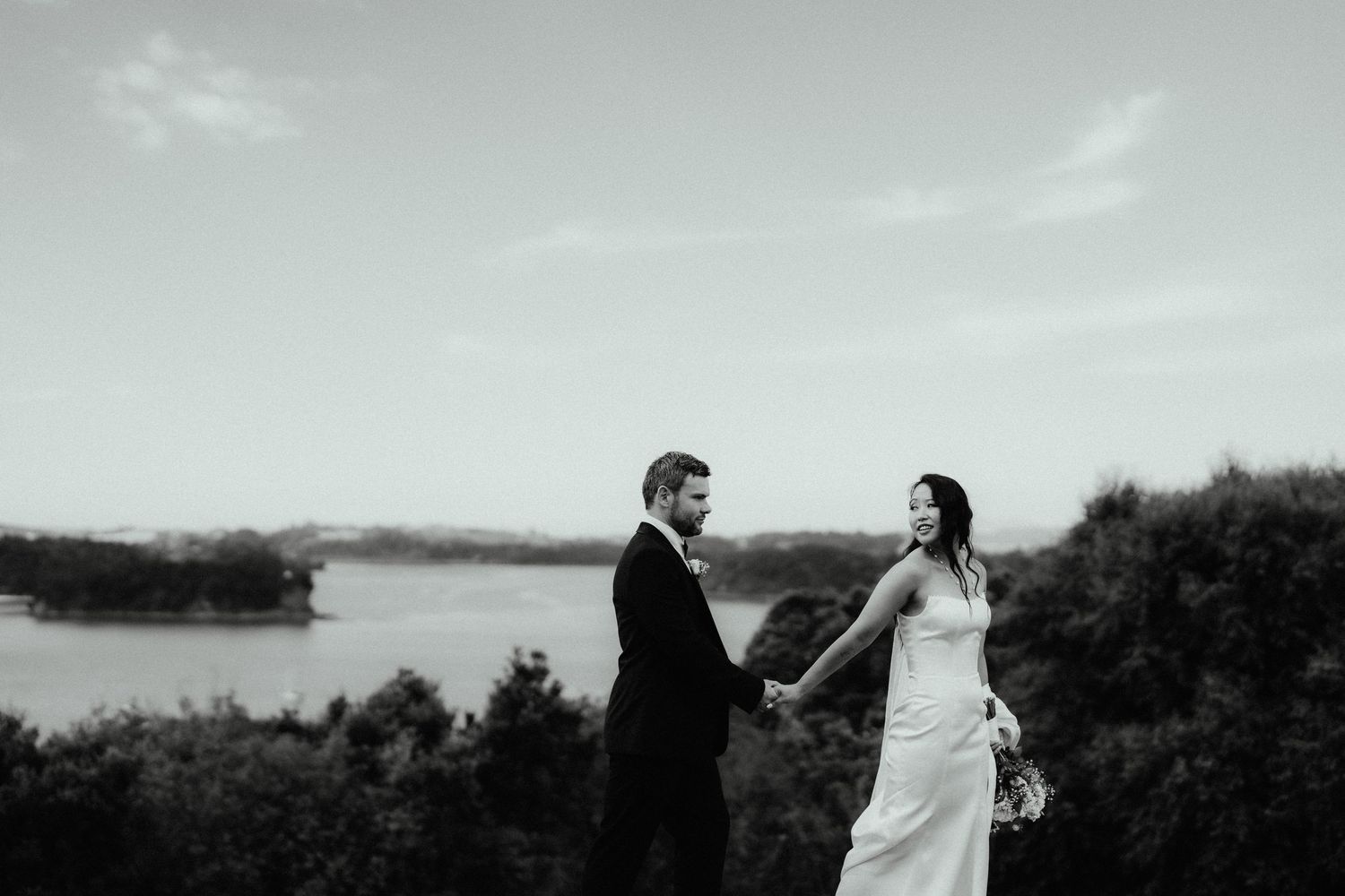 Bride and groom hold hands and smile at each other on a hilltop overlooking a calm bay in black and white.