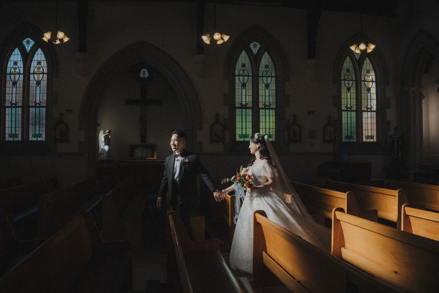 A bride and groom stand in a sunlit church aisle beneath stained glass windows and a wooden cross.