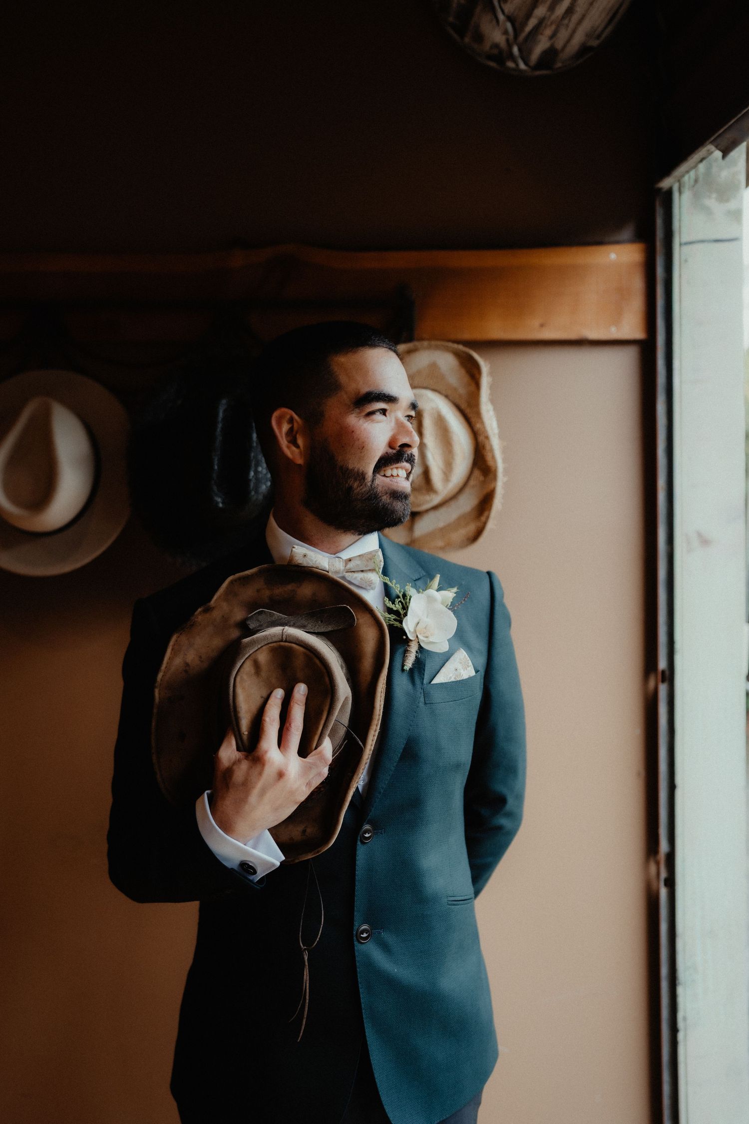 Smiling bearded groom in a teal suit with a white boutonniere holds a worn cowboy hat beside a window with hats on the wall.