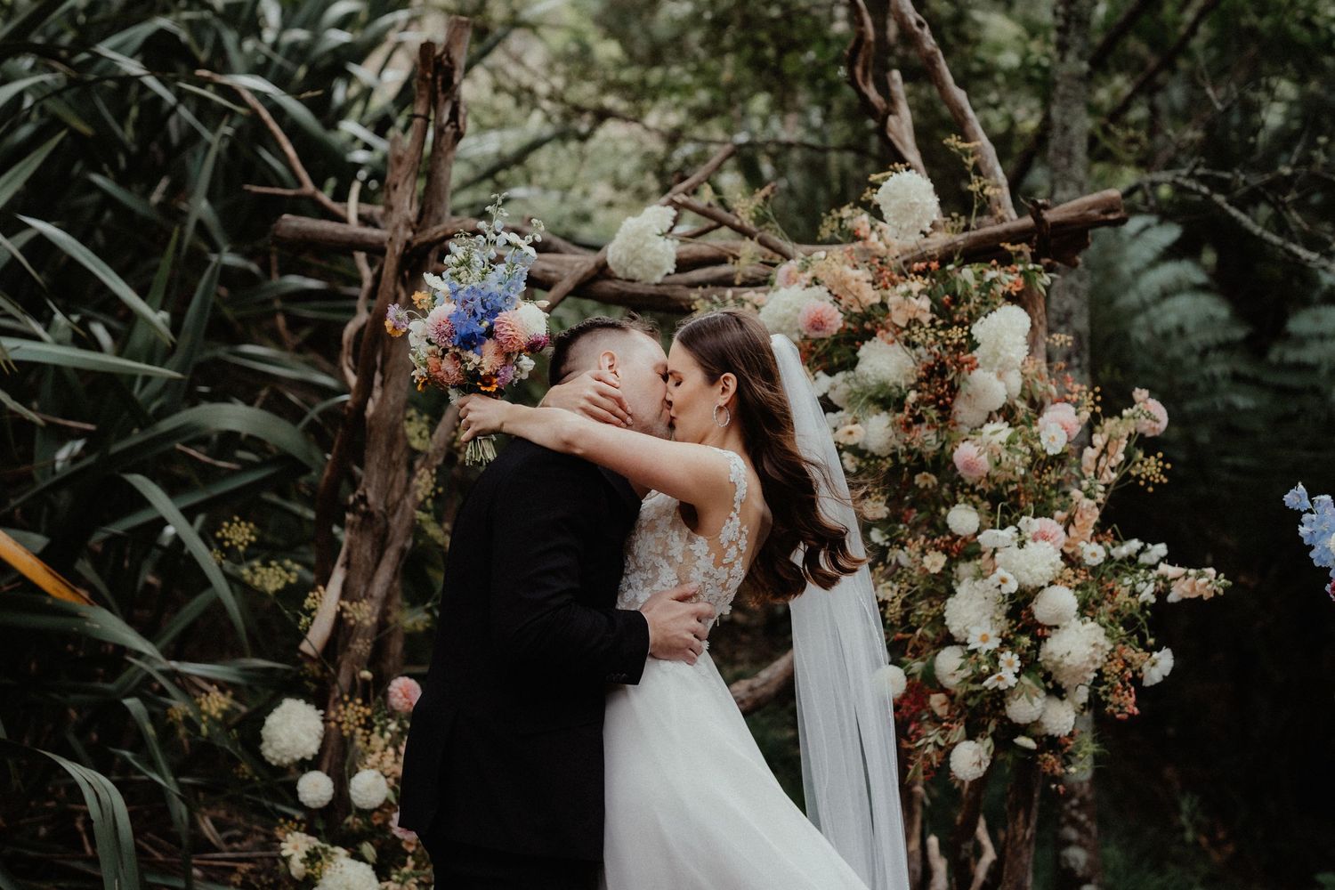 Bride and groom kiss beneath a rustic wooden arch decorated with white, pink and blue flowers in a lush garden.