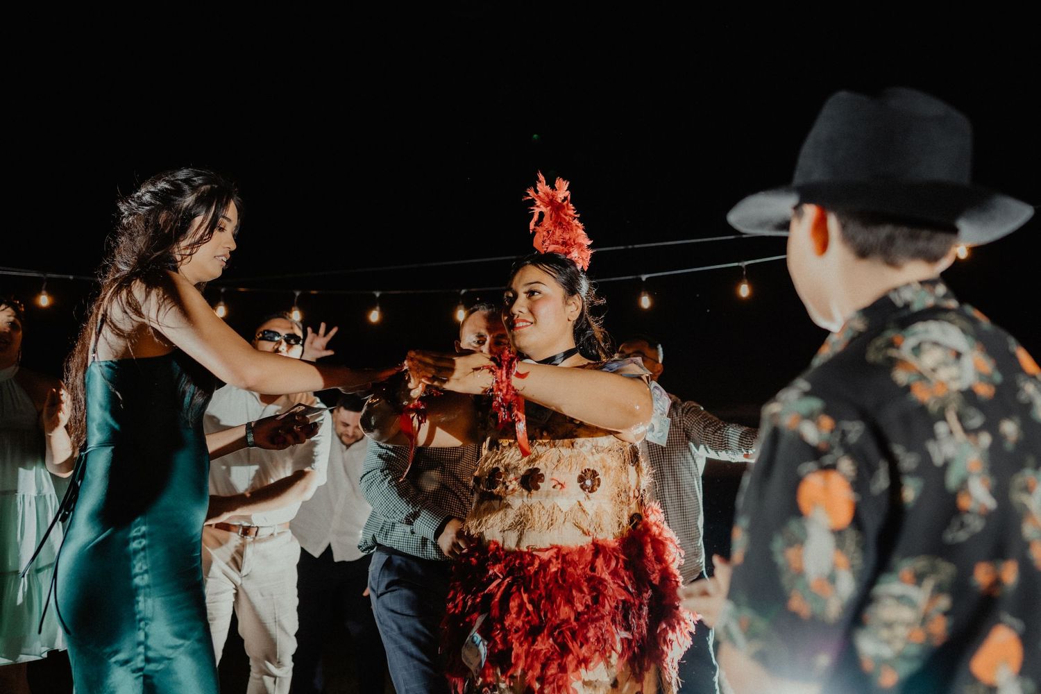 Wedding guests dancing at a night reception under string lights, with a woman in a red floral headpiece smiling at the center.
