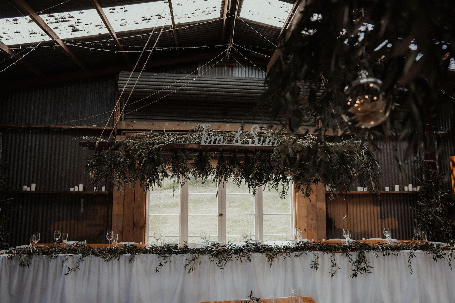 Rustic barn wedding reception venue with draped greenery, hanging lanterns, a name sign and white linen above the head table.