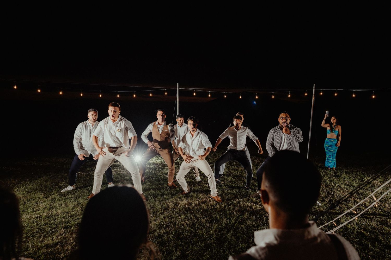 Groomsmen in white shirts crouch in a staggered line performing a haka-style dance at night on the grass beneath string lights.