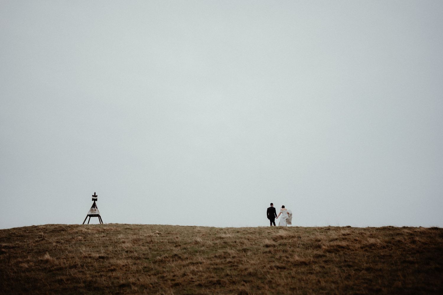 Tiny wedding couple walks hand in hand along a moody grassy hilltop beneath a vast grey sky near a small marker.