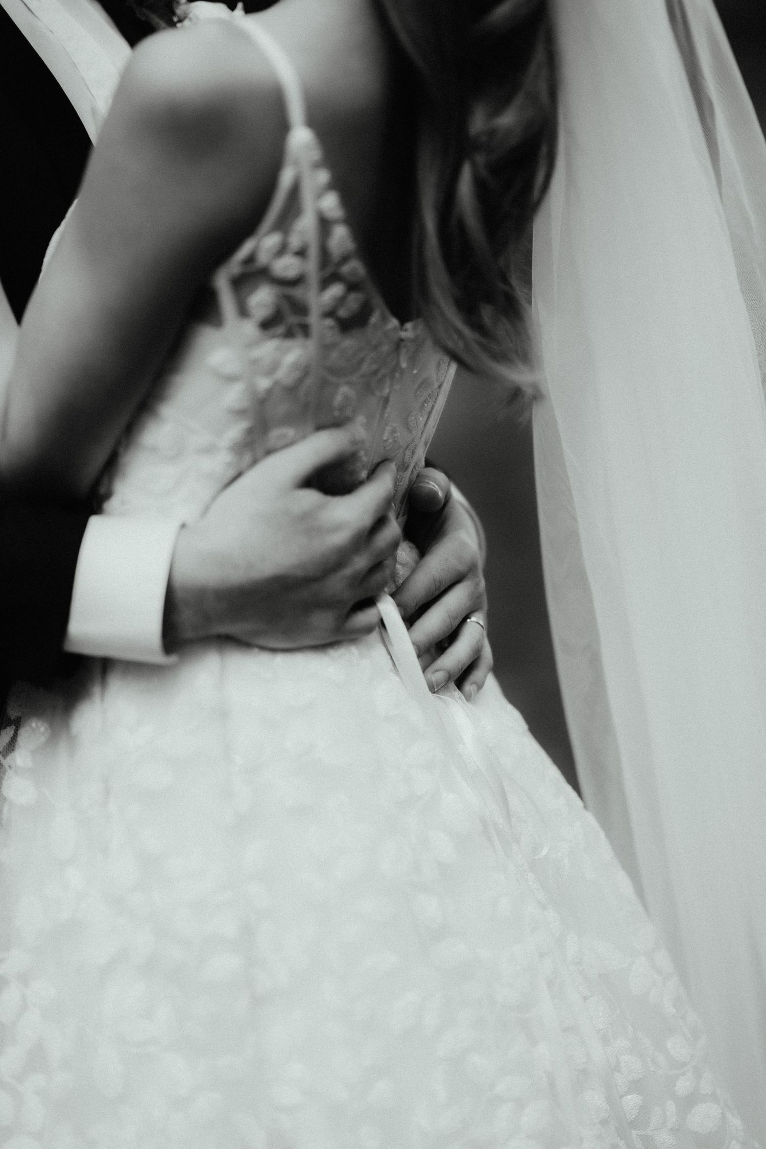 Black and white detail of a groom's hands resting on the lace back of a bride's wedding dress beside her veil.