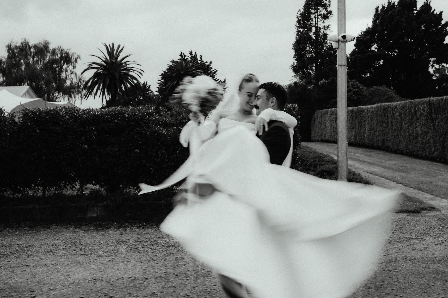 Black and white motion-blurred photo of a groom spinning his bride, her gown swirling, on a tree-lined path with palm trees.