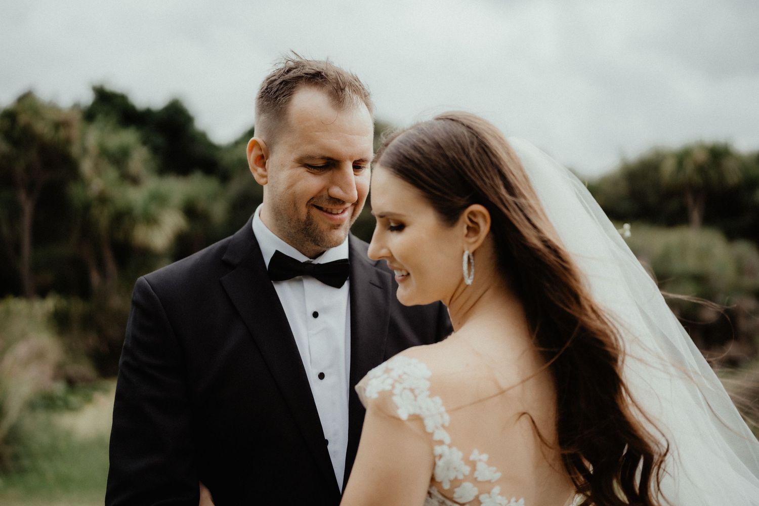 Smiling groom in a black tuxedo and bow tie gazes at his bride wearing a lace gown and long veil.