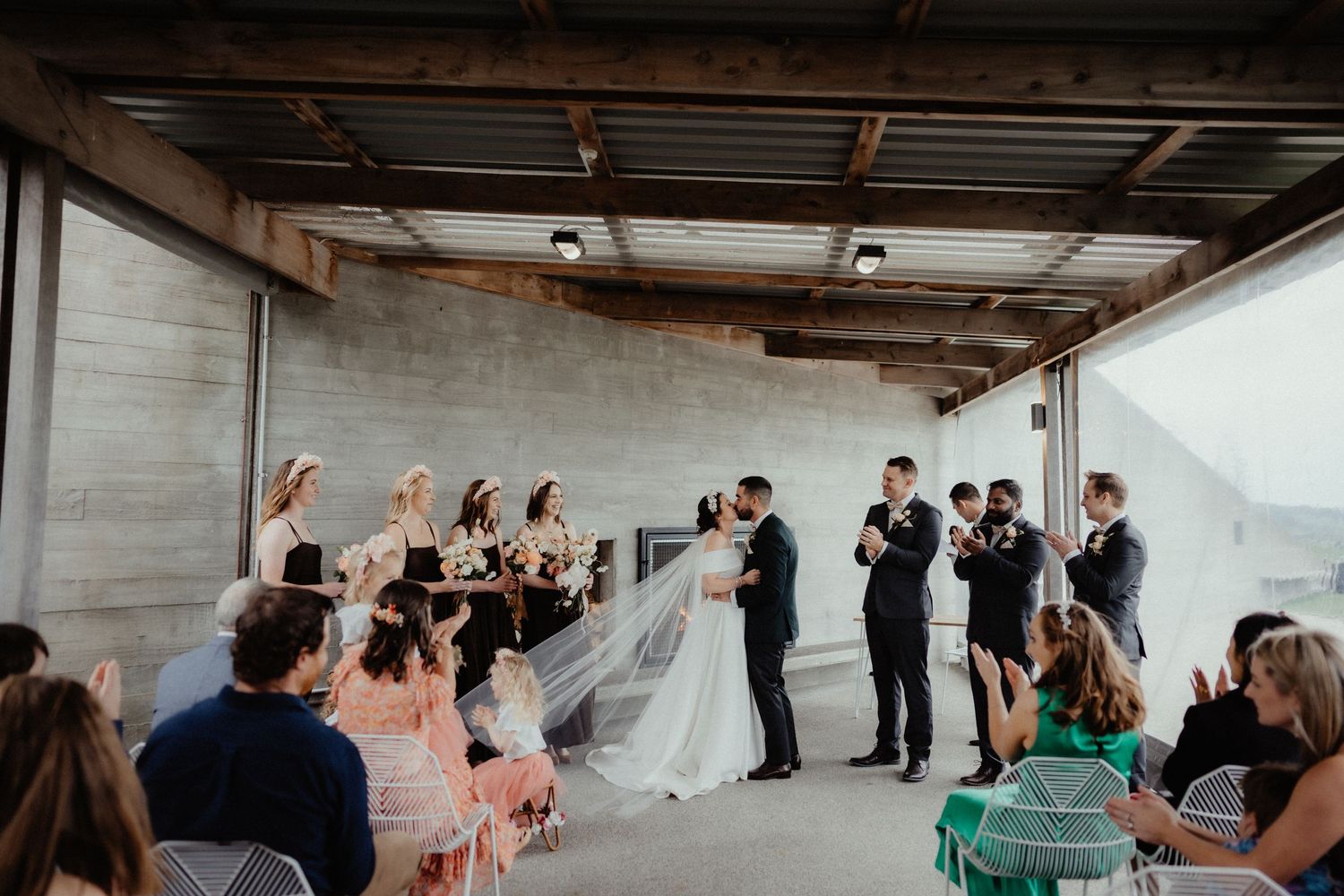 Bride and groom share a moment at the altar as bridesmaids, groomsmen and seated guests look on in a rustic venue.