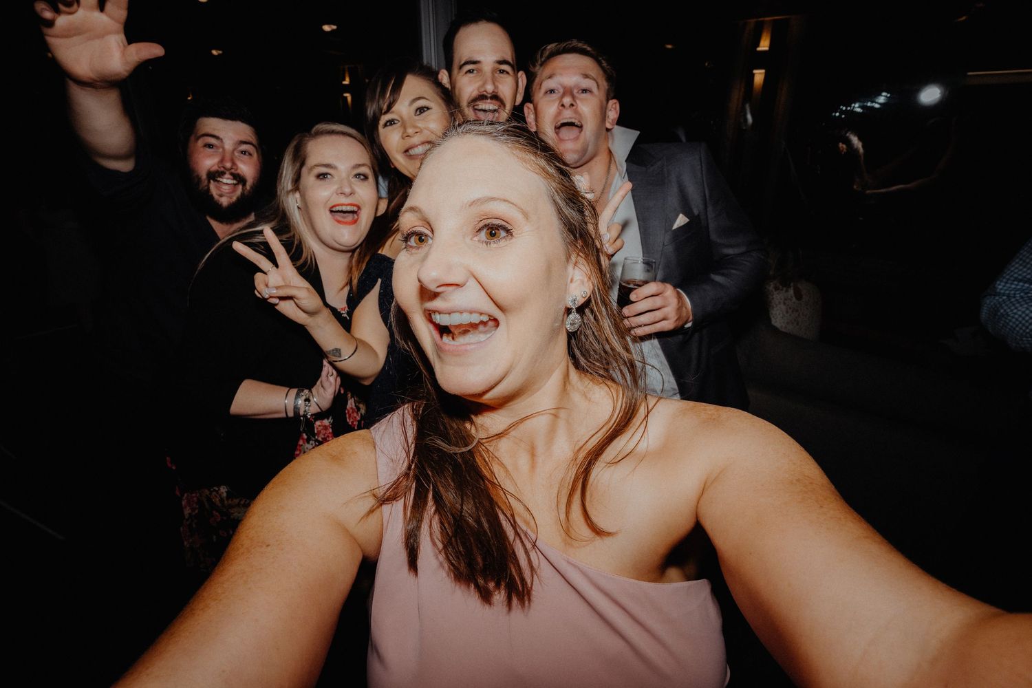 Wedding guests crowd close together laughing and posing for a lively selfie on the dance floor at night.