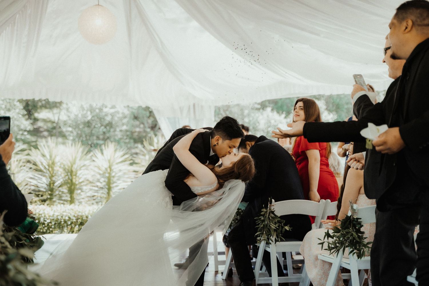 Groom dips the bride in a romantic kiss under a white draped tent as wedding guests cheer and take photos.