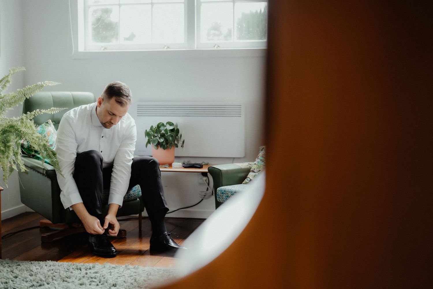 Groom in white shirt and dark trousers sits in a green armchair tying his shoes by a bright window before the wedding.