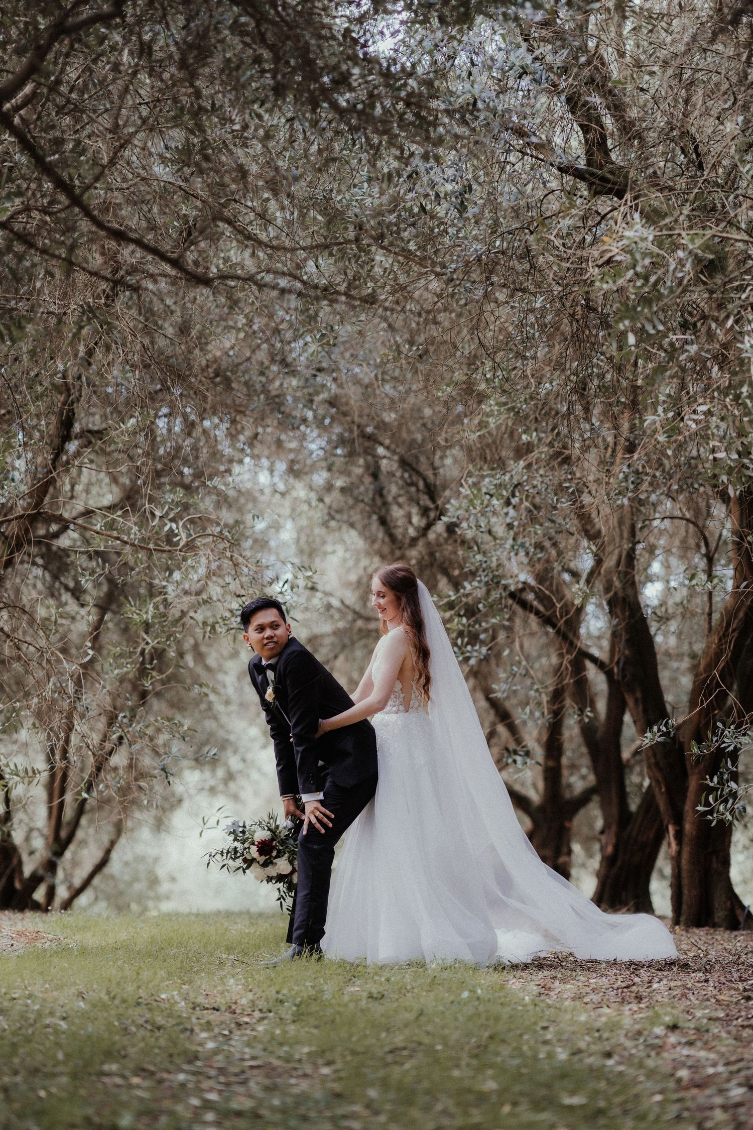 Bride in a white gown and long veil playfully stands behind a groom in a black suit beneath olive trees.