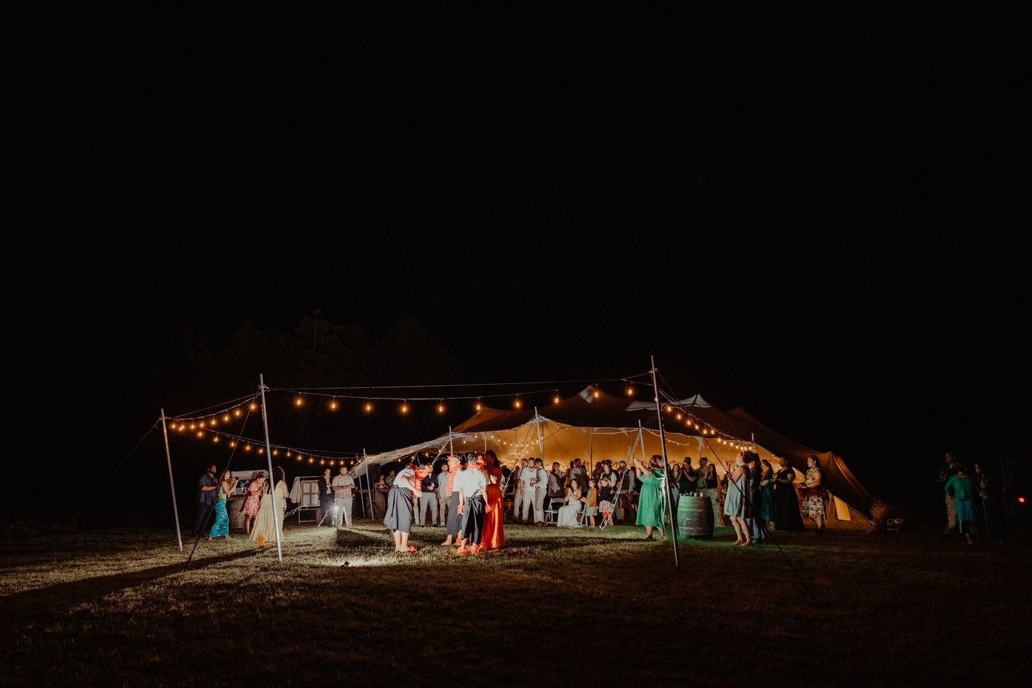 Wedding guests gather under a lit tent strung with festive lights in a dark field during an evening reception.