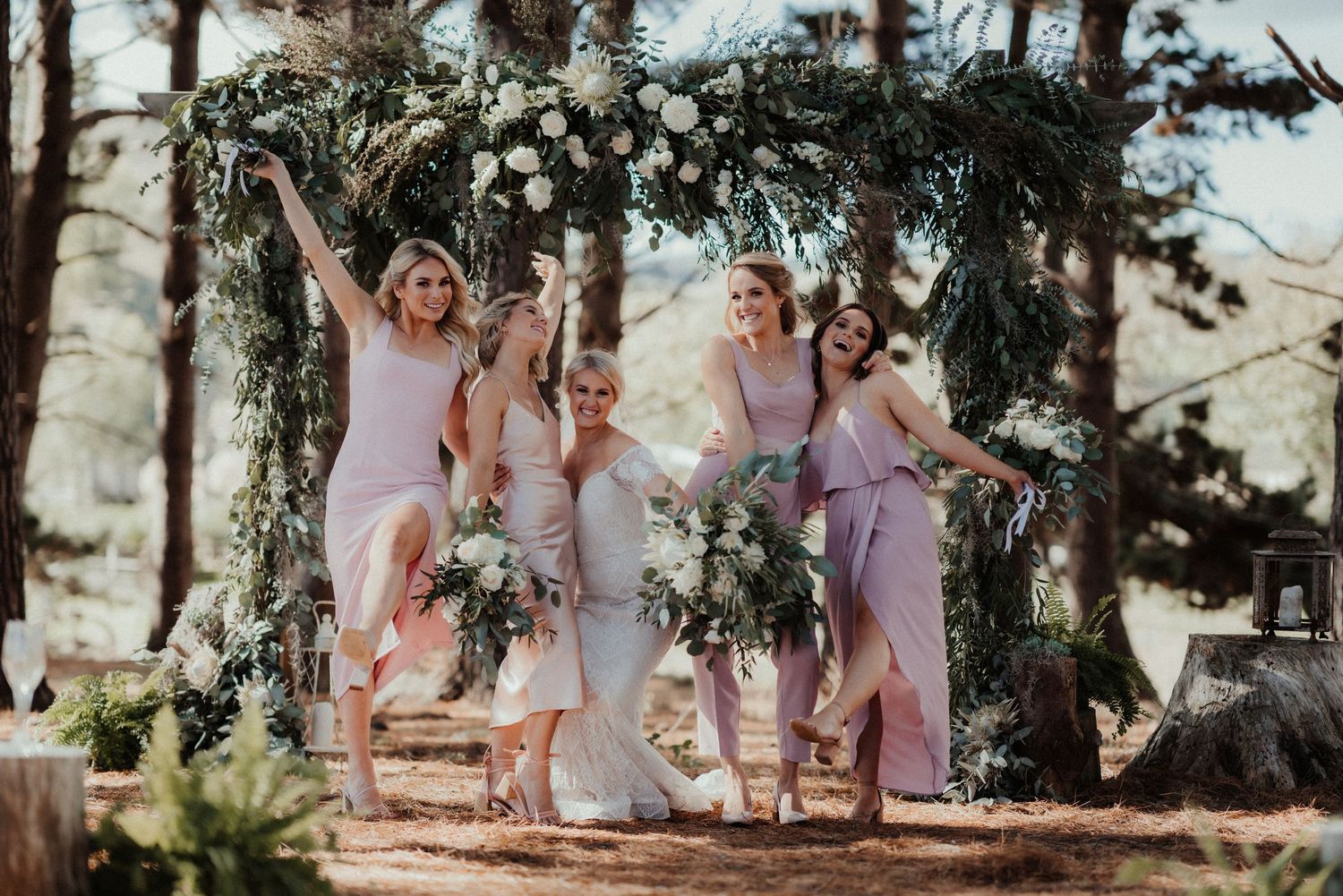 Bridesmaids in dusty mauve dresses laugh and pose with a bride holding bouquets in front of a floral arch in a pine forest.