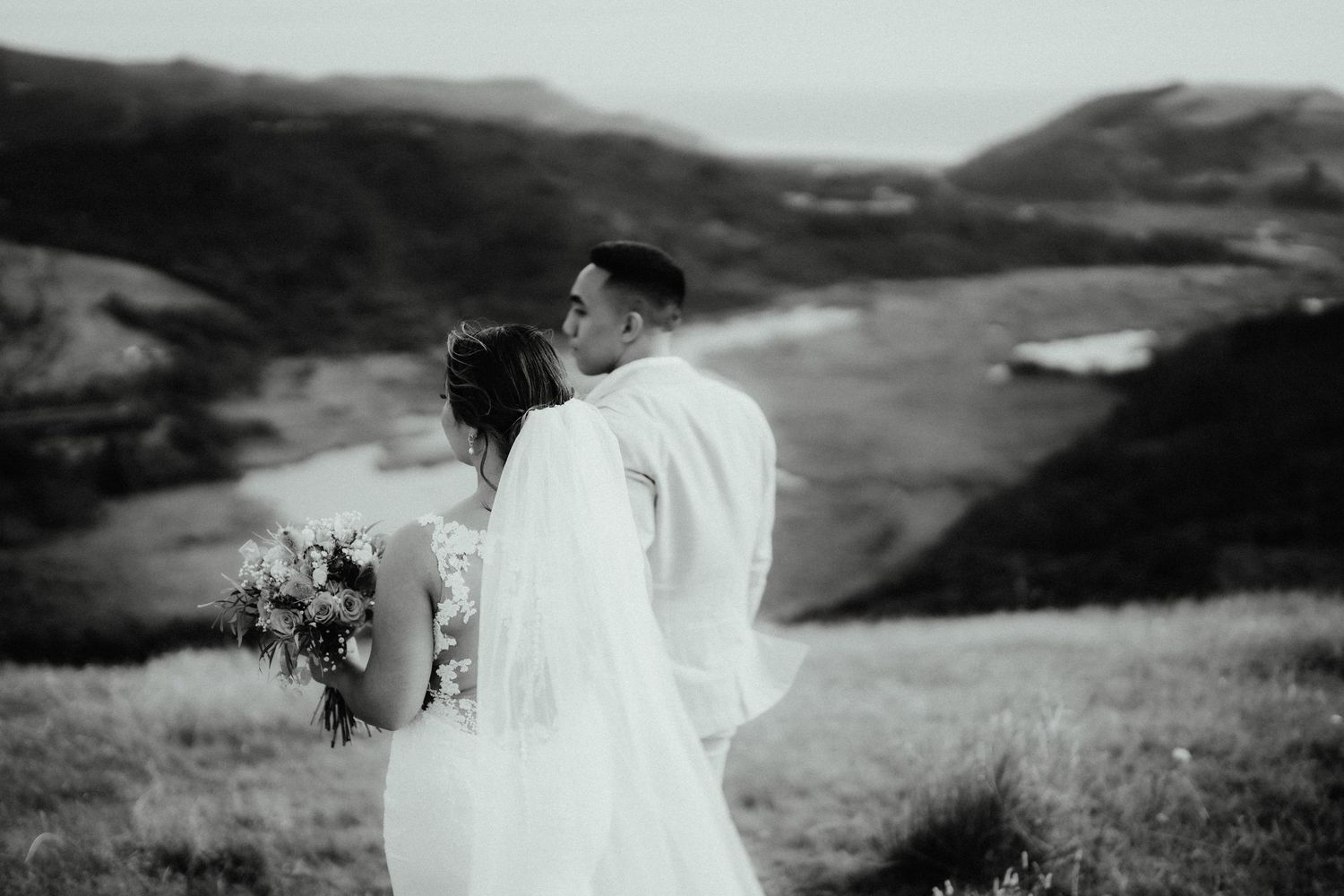 Black and white photo of a bride in a long veil holding a bouquet while embracing her groom on a grassy hilltop overlooking the coast.