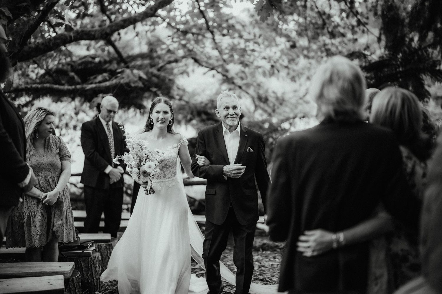 Black and white photo of a father walking his smiling bride down the aisle as guests look on at an outdoor wedding.