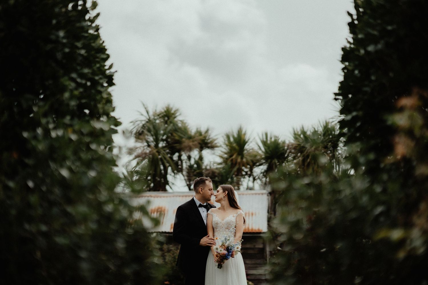 Bride and groom embrace on a garden path framed by tall hedges with palm trees and a cloudy sky behind them.