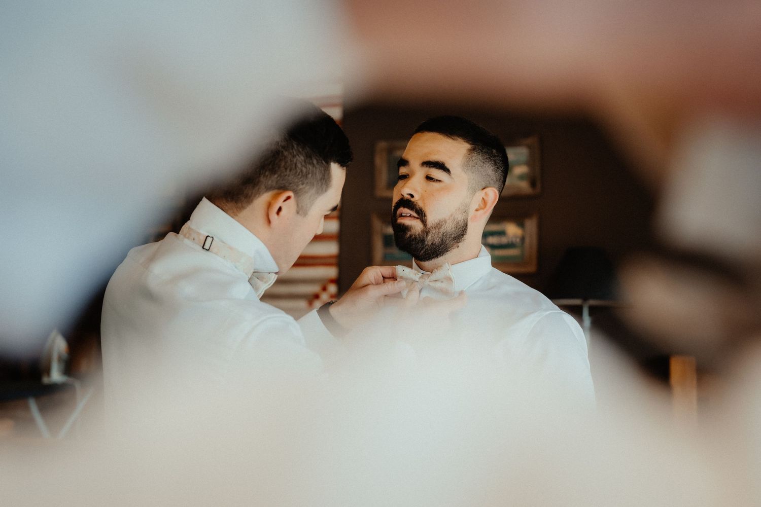 Groom with a beard has his bow tie adjusted by a groomsman while getting ready for the wedding.