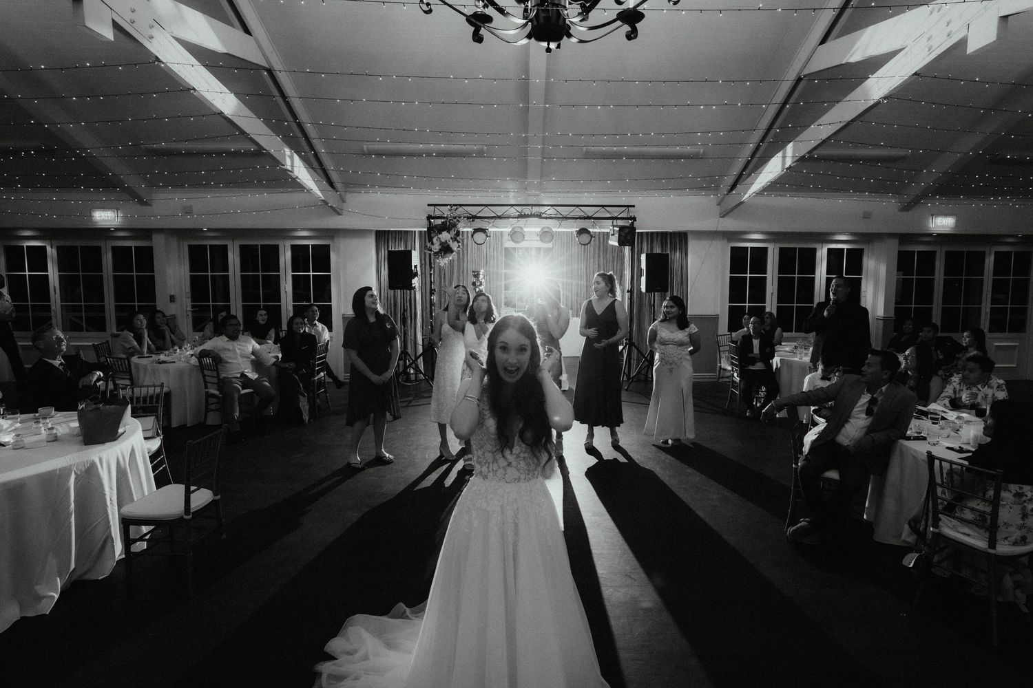 Black and white photo of a bride in a white gown standing on the dance floor as guests look on at a wedding reception.